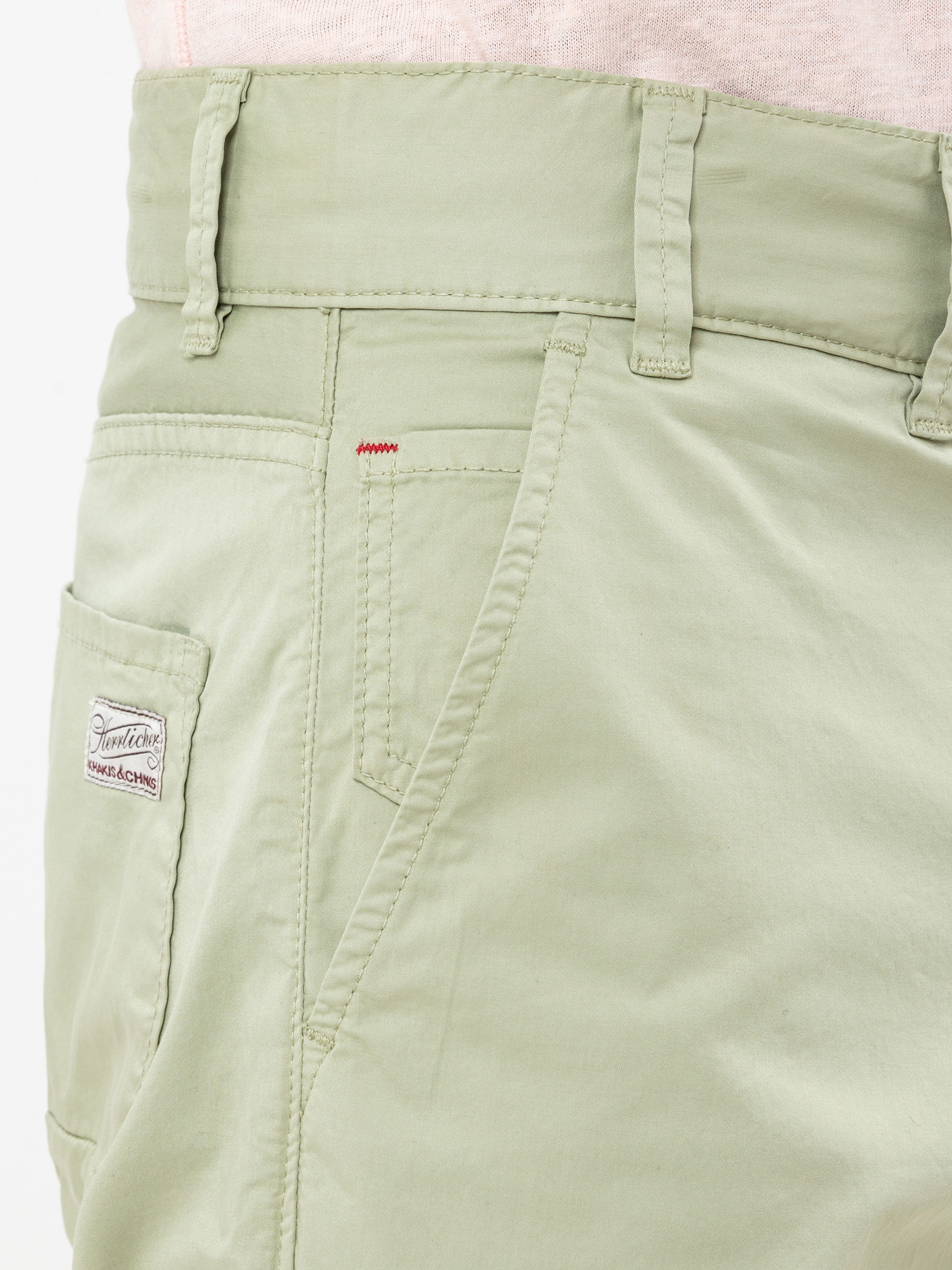 Nahaufnahme einer hellgrünen Hose mit einer Münztasche, Gürtelschlaufen, einer Seitentasche und einem kleinen aufgenähten Etikett mit der Aufschrift Trademark auf der Gesäßtasche; rote Akzentnähte über der Haupttasche.