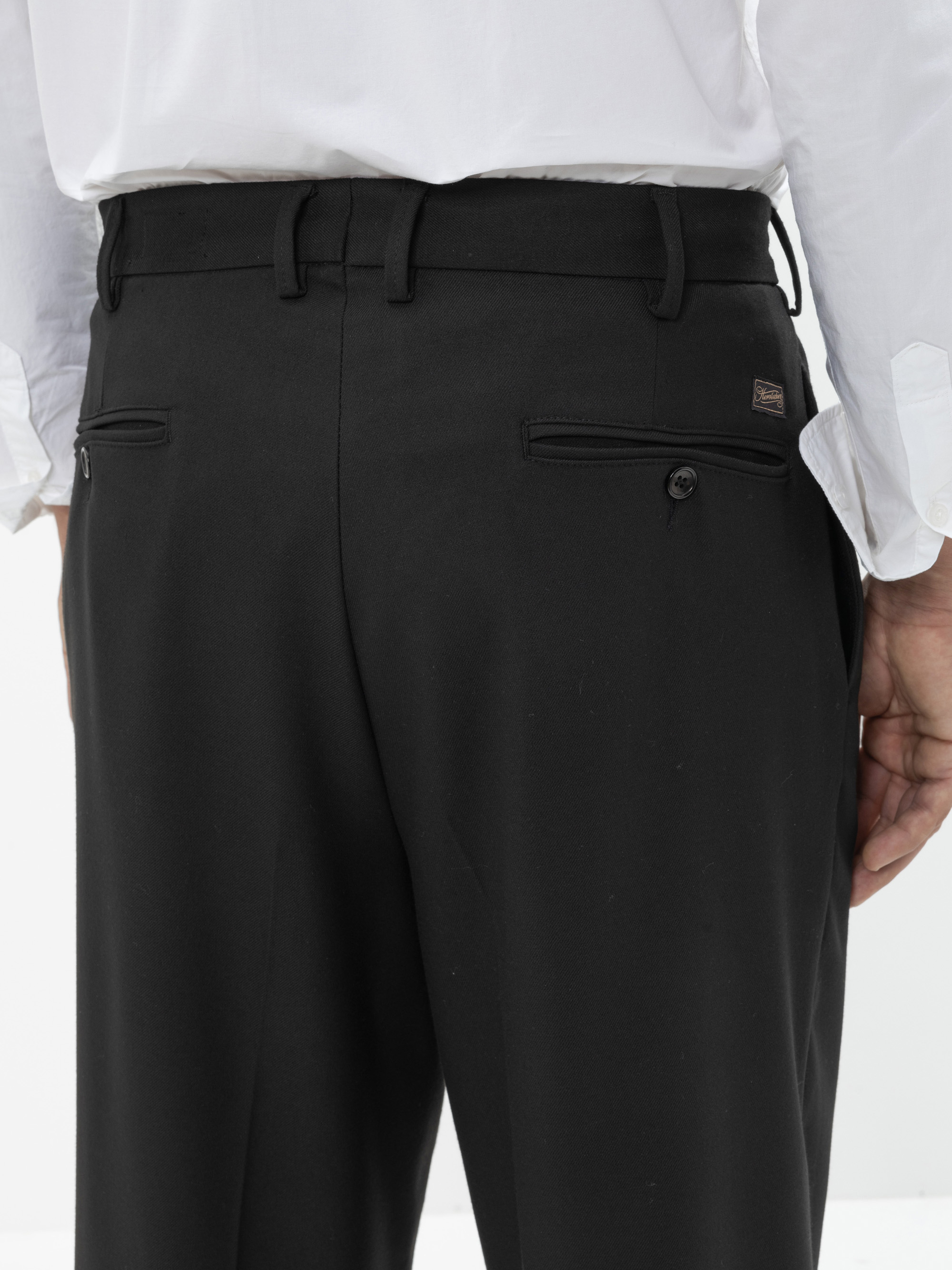Eine Person trägt ein weißes Hemd und eine schwarze Anzughose, von hinten gesehen. Die Hose hat Gürtelschlaufen und zwei Gesäßtaschen, eine mit einem Knopf und eine mit einem Reißverschluss.