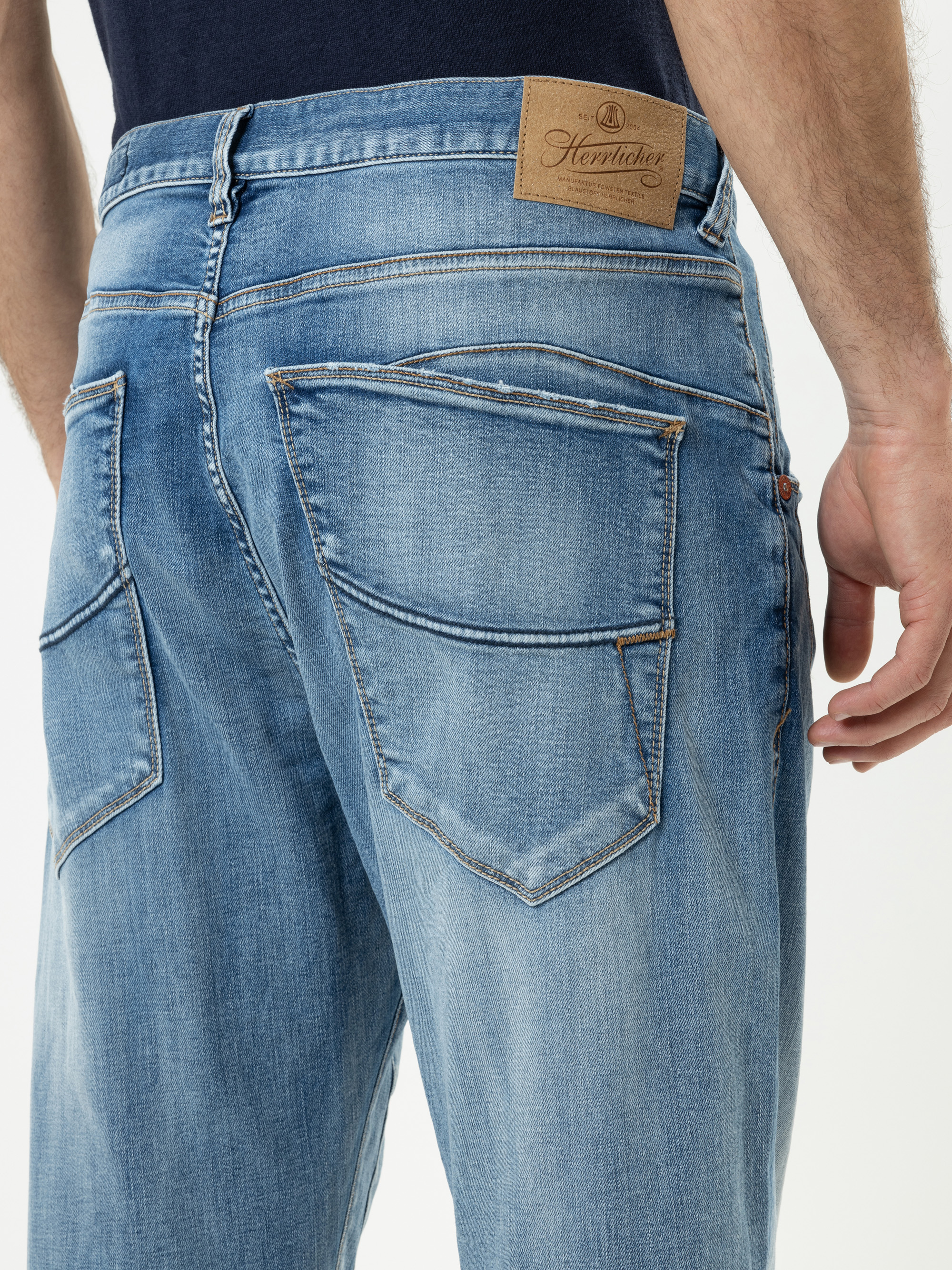 Eine Person in hellblauer Jeans und schwarzem Hemd wird von hinten gezeigt. Die Jeans hat große Gesäßtaschen mit orangefarbenen Nähten und einen braunen Markenaufnäher aus Leder am Bund.
