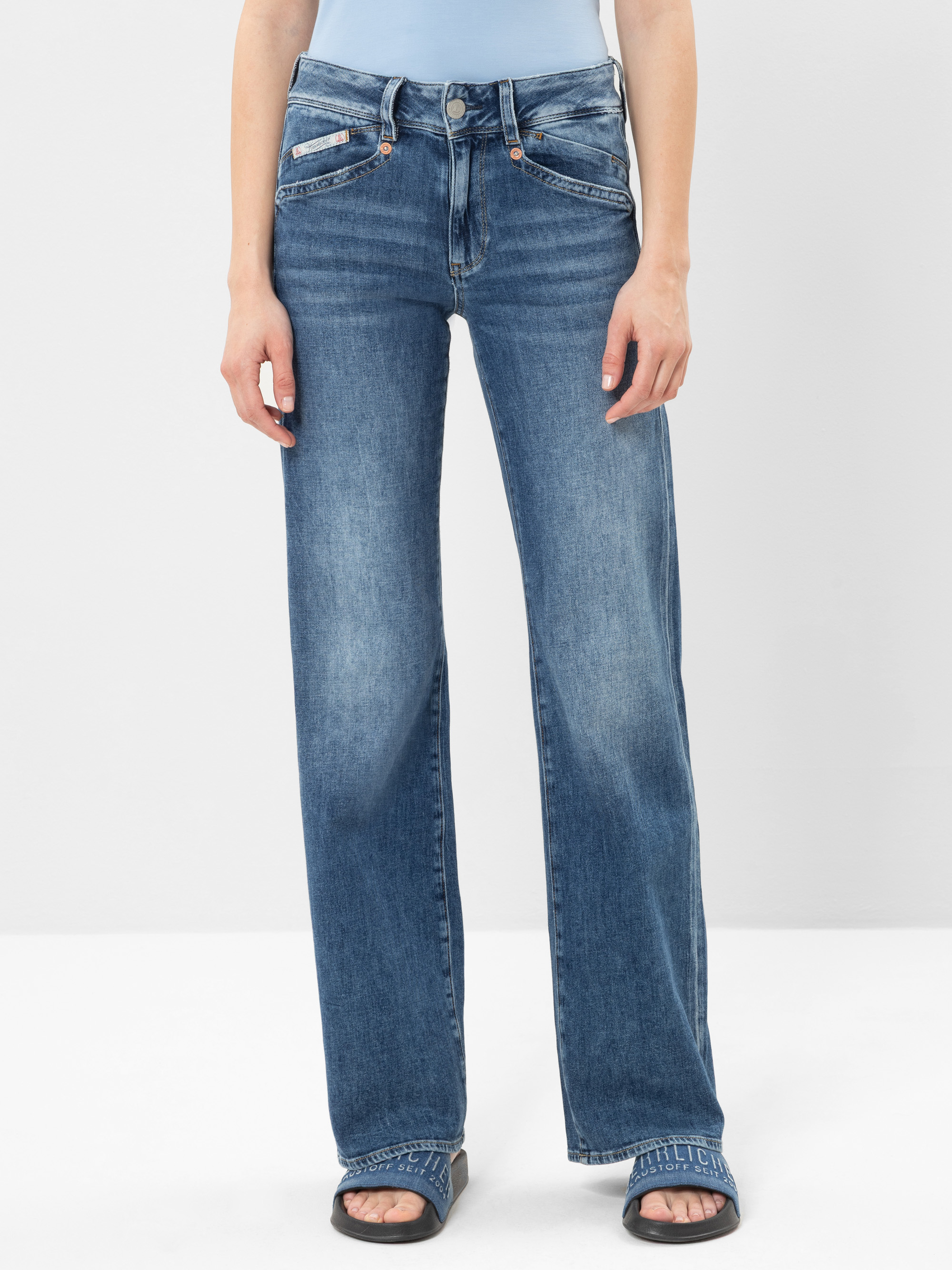 Person in blauen Jeans mit geradem Bein und schwarzen Sandalen, stehend vor einem schlichten weißen Hintergrund. Nur der untere Teil des Oberkörpers und die Beine sind sichtbar.