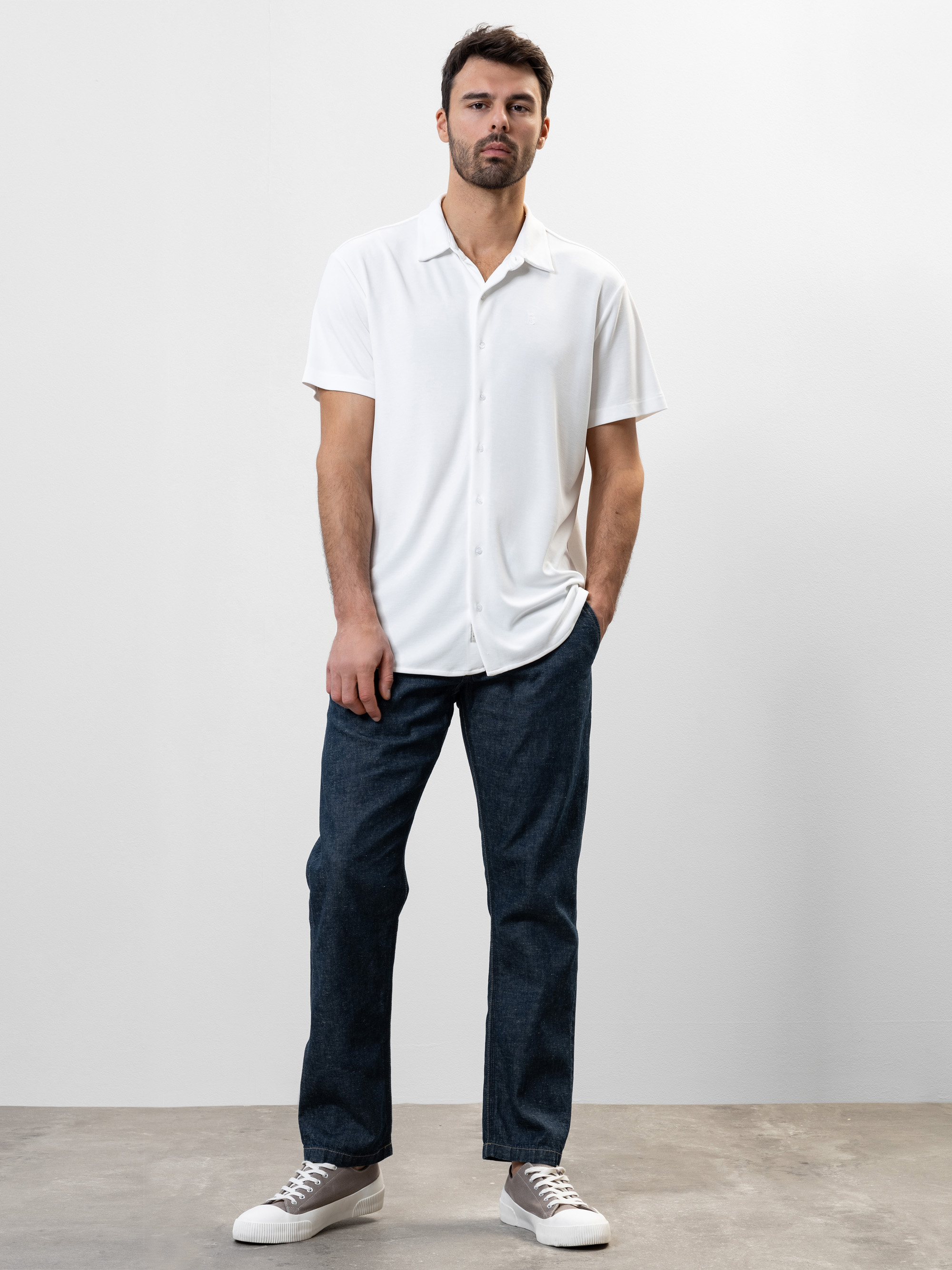 Ein Mann mit kurzen dunklen Haaren steht vor einem schlichten hellen Hintergrund. Er trägt ein weißes, kurzärmeliges Button-up-Hemd, dunkelblaue Jeans und weiße Turnschuhe und hat eine Hand in der Tasche.