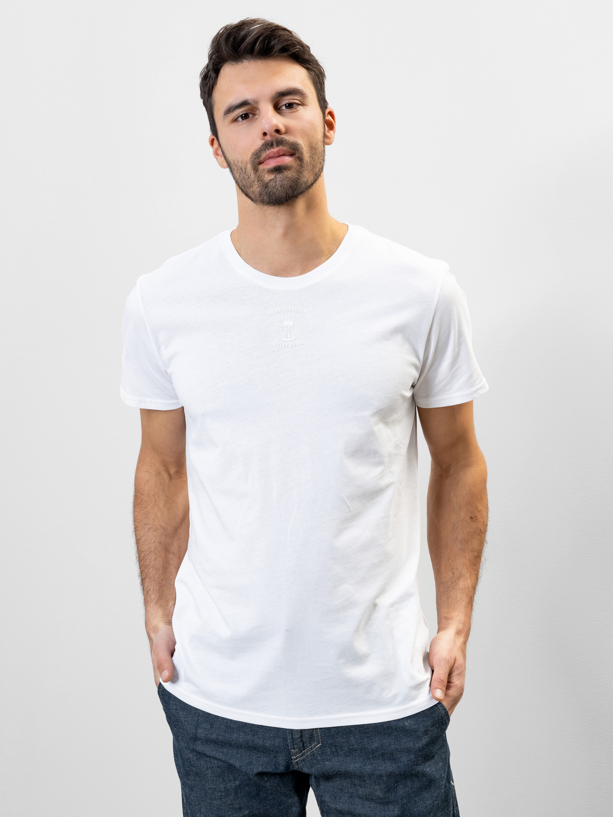 Ein Mann mit kurzen dunklen Haaren und einem Bart steht vor einem schlichten hellen Hintergrund. Er trägt ein schlichtes weißes T-Shirt und dunkle Jeans, hat die Hände in den Taschen und schaut ruhig in die Kamera.