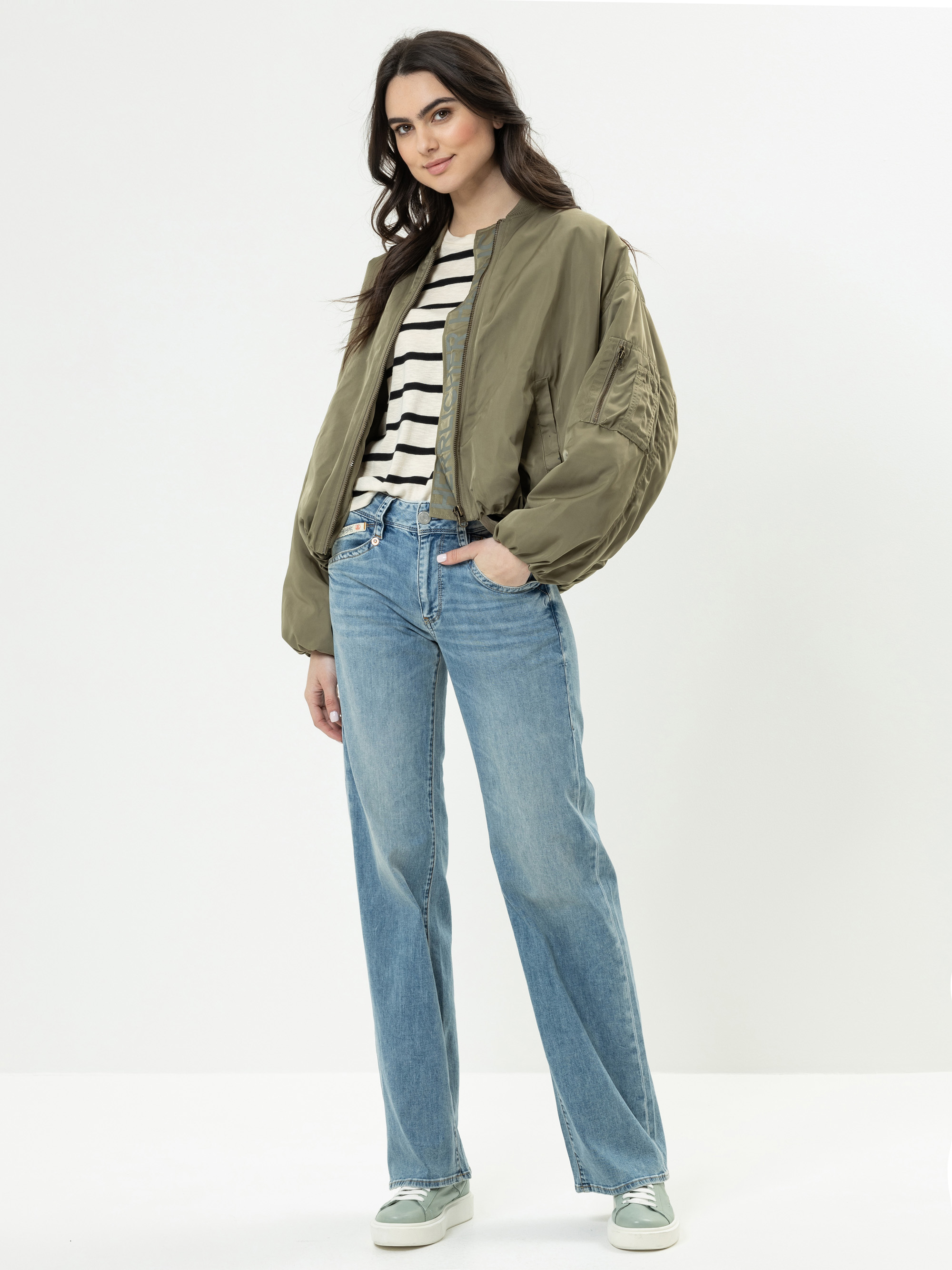 Eine Frau steht vor einem schlichten Hintergrund und trägt eine khakifarbene Bomberjacke, ein schwarz-weiß gestreiftes Hemd, blaue High-Waisted-Jeans und helle Turnschuhe. Sie hat langes, gewelltes dunkles Haar und eine entspannte, selbstbewusste Pose.