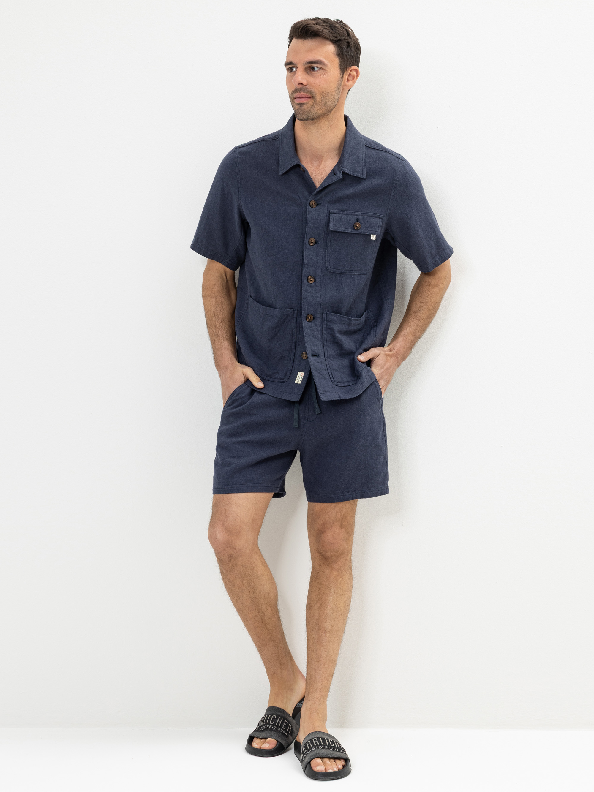 Ein Mann steht vor einem weißen Hintergrund und trägt ein marineblaues, kurzärmeliges Button-up-Hemd mit Vordertaschen, dazu passende marineblaue Shorts und schwarze Slide-Sandalen. Seine Hände befinden sich in den Taschen und er blickt leicht zur Sei