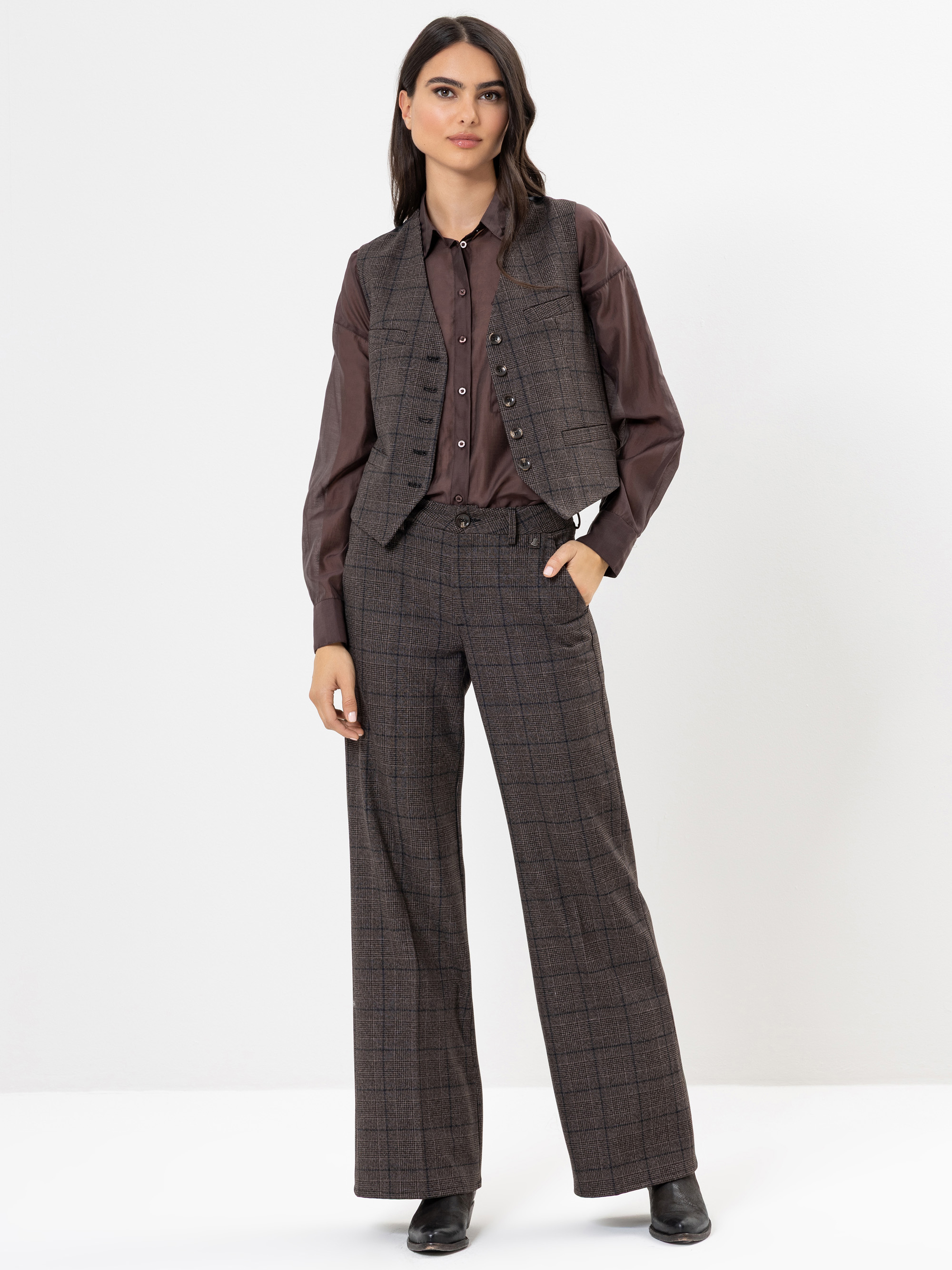 Eine Frau steht vor einem weißen Hintergrund und trägt ein braunes Button-up-Hemd, eine passende karierte Weste und eine weit geschnittene karierte Hose. Sie hat eine Hand in ihrer Tasche und trägt schwarze Schuhe.