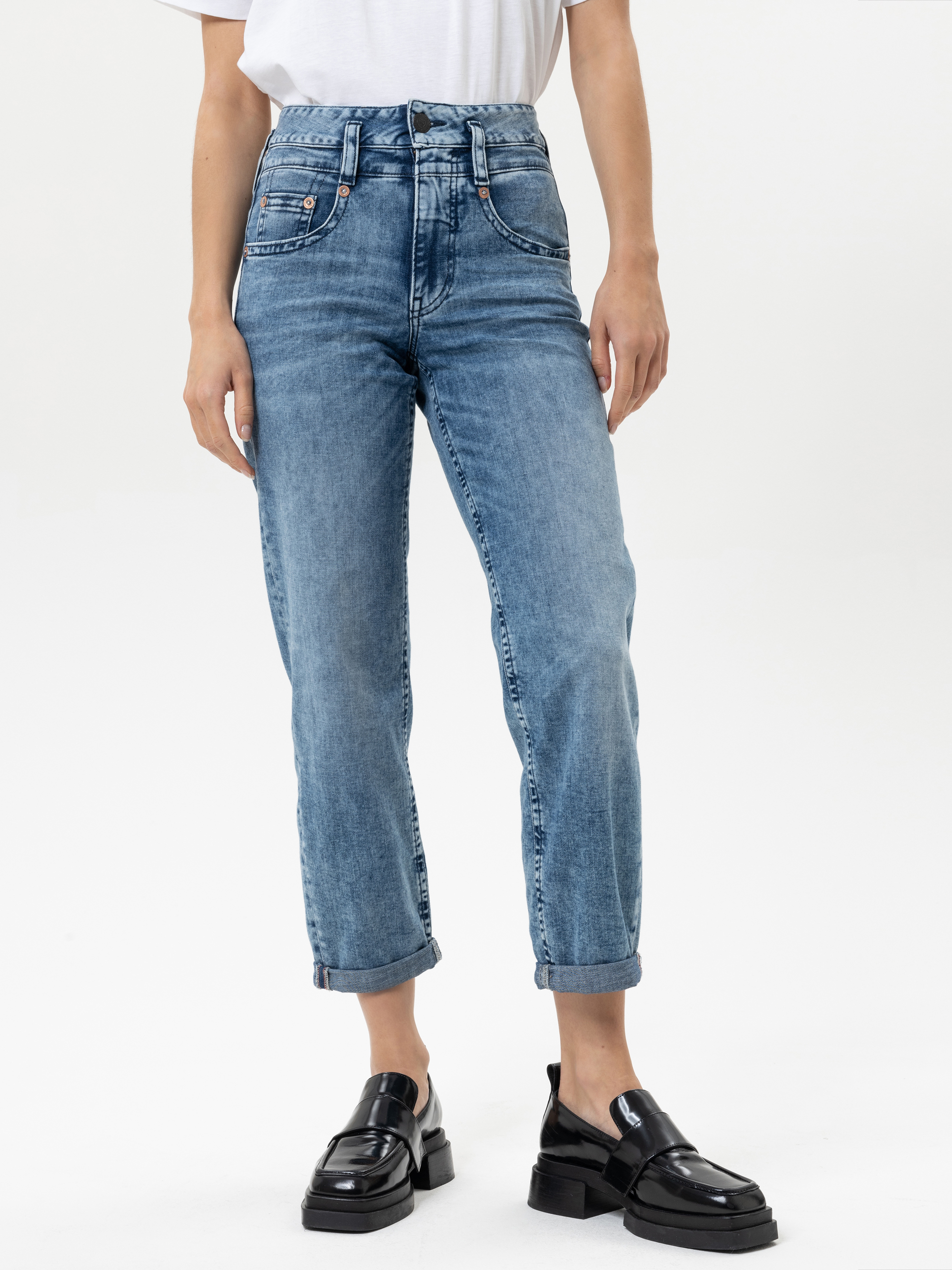 Eine Person, die eine hochgekrempelte Bluejeans, ein weißes T-Shirt und schwarze Slipper trägt, steht vor einem schlichten weißen Hintergrund. Nur der untere Teil des Oberkörpers und die Beine sind sichtbar.