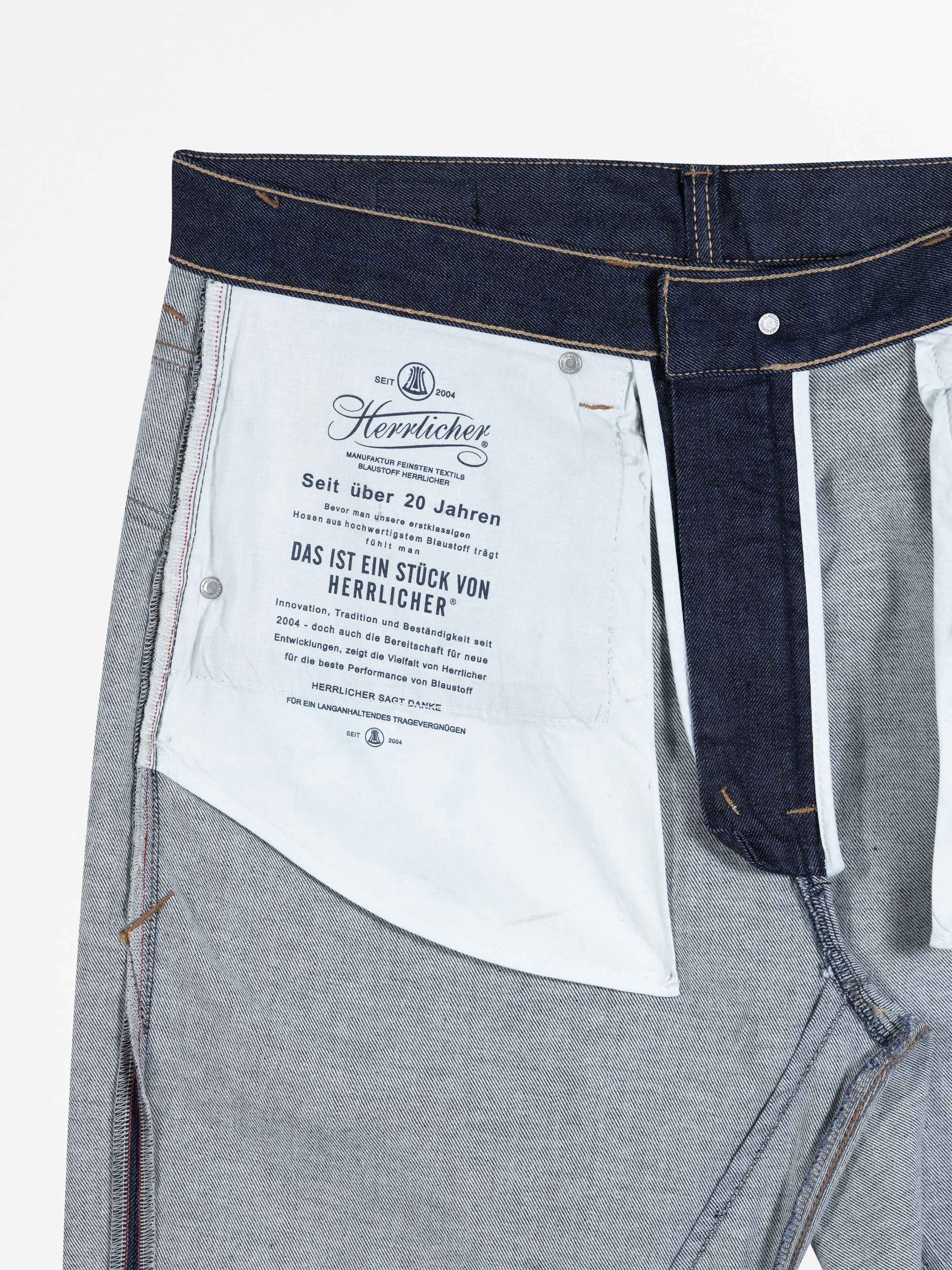 Nahaufnahme der Innenseite einer Jeans, die ein weißes Taschenfutter mit aufgedrucktem deutschen Text und Branding von Herrlicher zeigt. Der Denim und die Nähte sind deutlich sichtbar.
