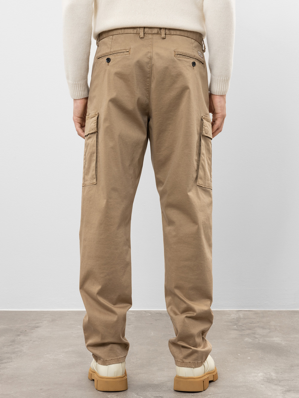 Eine Person, die eine hellbraune Cargohose, einen cremefarbenen langärmeligen Pullover und beigefarbene Schuhe trägt, steht mit dem Gesicht zur Kamera vor einem schlichten, hellen Hintergrund.