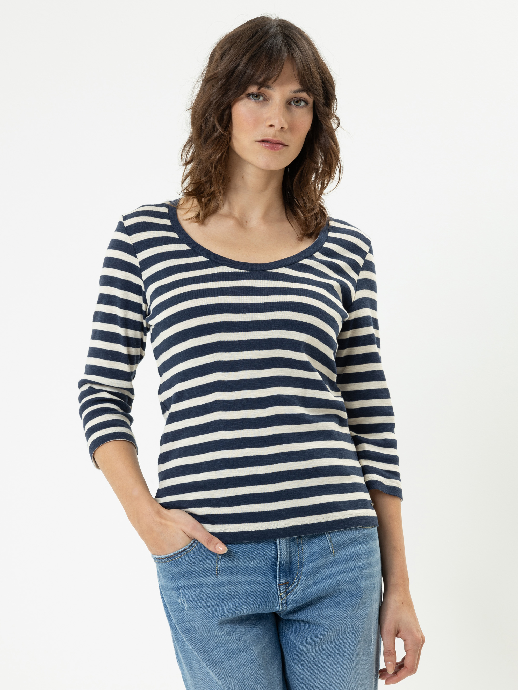 Eine Frau mit gewelltem braunem Haar trägt ein marineblau-weiß gestreiftes langärmeliges Hemd und blaue Jeans. Sie steht vor einem schlichten weißen Hintergrund und hat eine Hand in der Tasche.