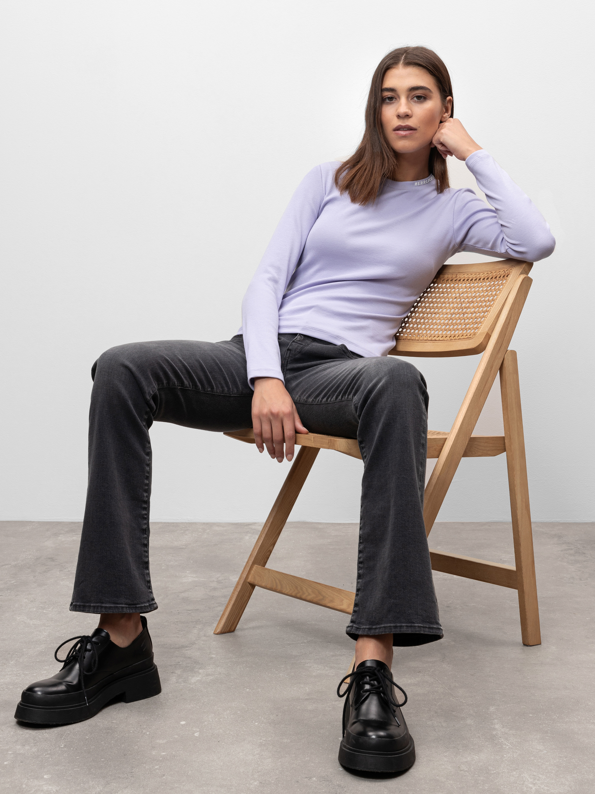 Eine Frau in einem hellvioletten langärmeligen Oberteil und dunklen Schlagjeans sitzt lässig auf einem Holzstuhl mit geflochtener Rückenlehne, einen Arm auf den Stuhl gestützt, den anderen auf ihr Knie, vor einem schlichten grauen Hintergrund.