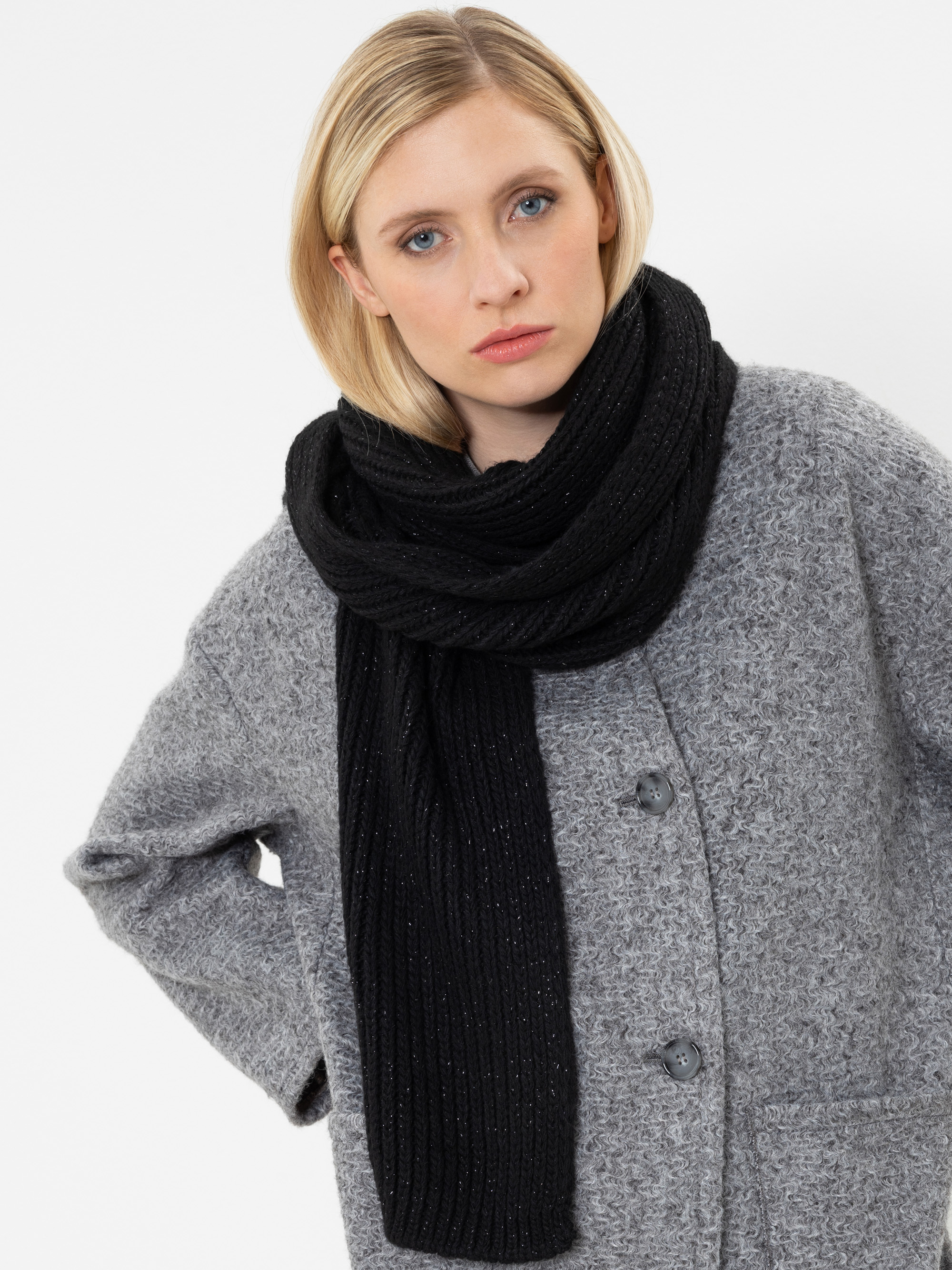 Eine Frau mit blonden Haaren, die einen grauen Mantel mit Knöpfen und einen dicken schwarzen Strickschal trägt, steht vor einem schlichten weißen Hintergrund und blickt mit ausdruckslosem Gesicht in die Kamera.
