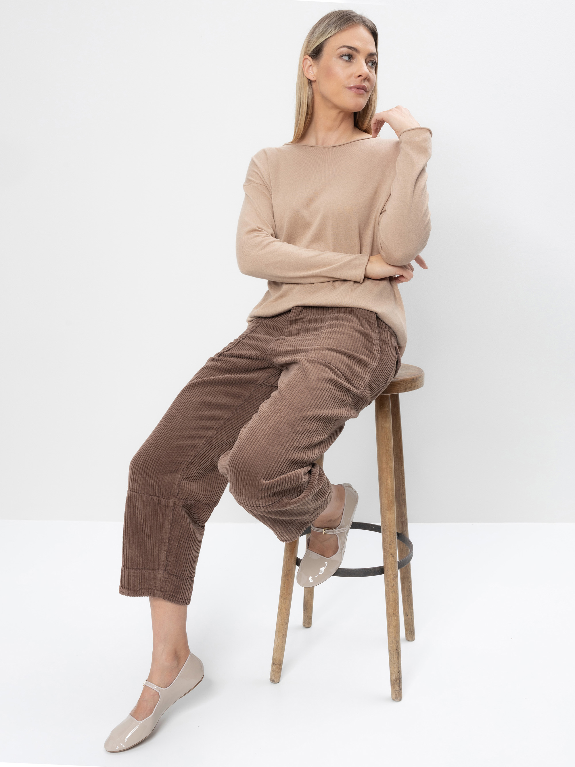 Eine Frau in einem beigefarbenen langärmeligen Oberteil und einer braunen Cordhose sitzt auf einem Holzhocker vor einem schlichten weißen Hintergrund und blickt zur Seite, wobei ein Arm auf ihrem angehobenen Knie ruht. Sie trägt hellbeigefarbene flache