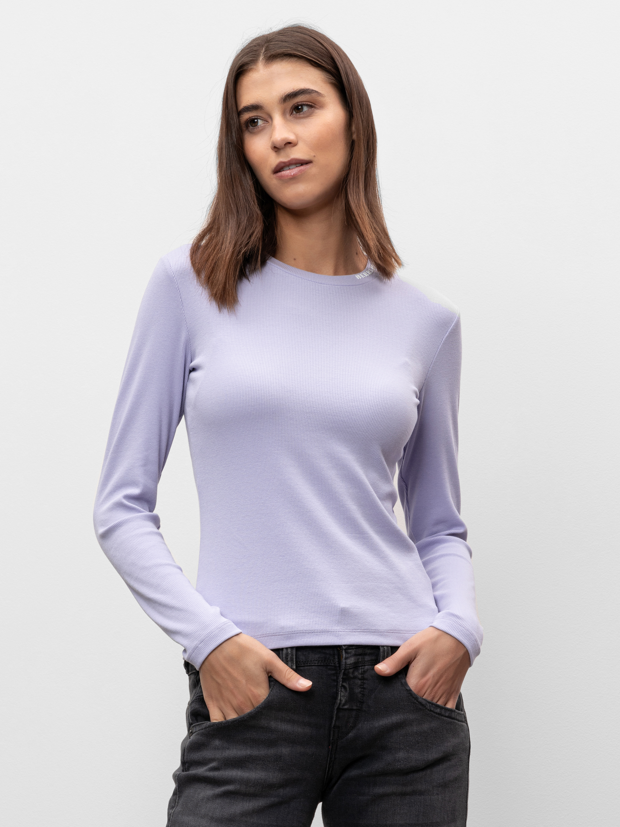 Eine Frau mit glattem braunem Haar, die ein hellviolettes langärmeliges Oberteil und schwarze Jeans trägt, steht vor einem schlichten hellen Hintergrund und schaut leicht zur Seite, die Hände in den Taschen.