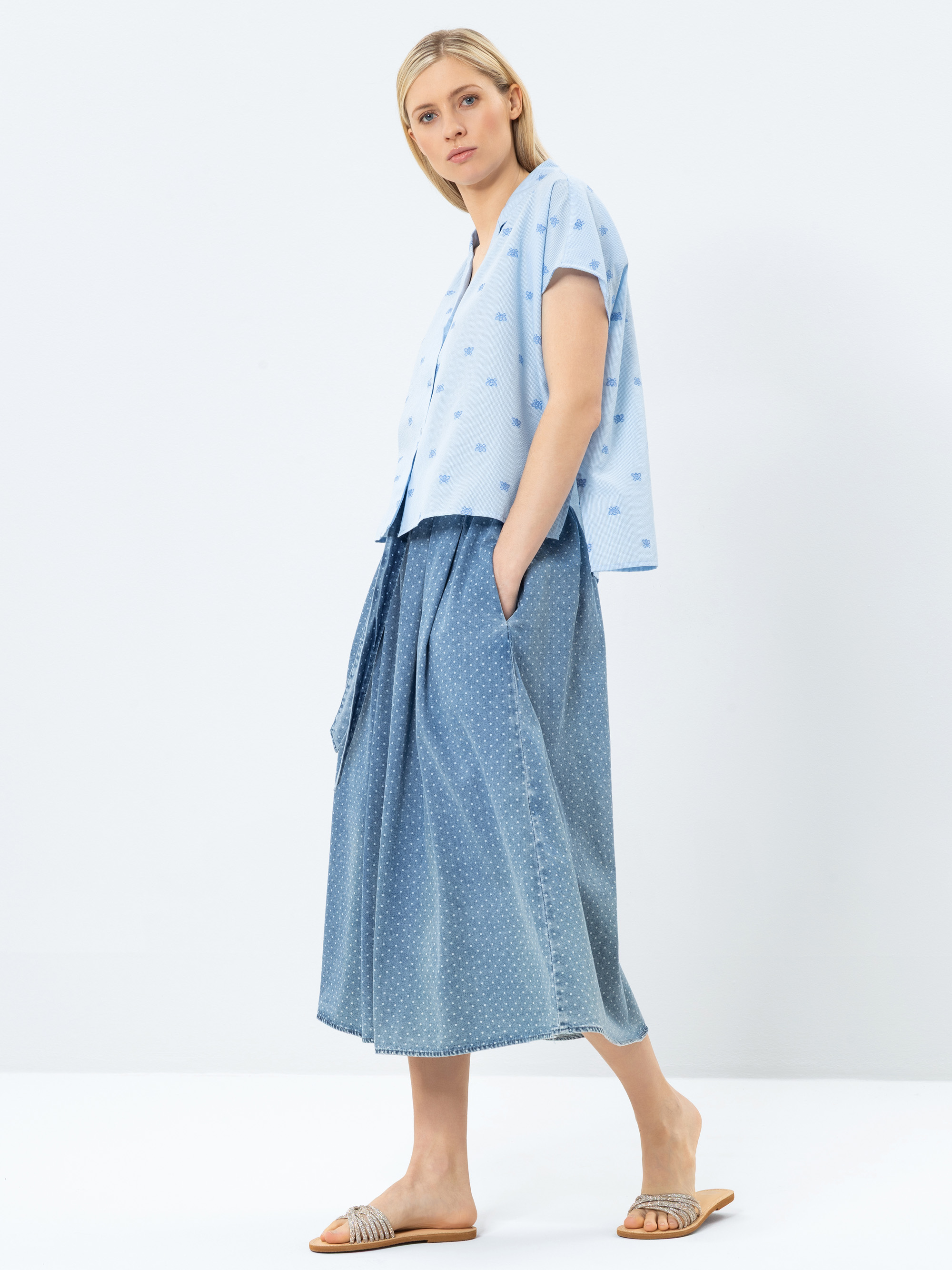 Eine Frau steht seitlich mit den Händen in den Taschen. Sie trägt ein hellblaues, kurzärmeliges Button-up-Hemd, einen fließenden blauen Midirock mit einem dezenten Muster und flache Sandalen. Der Hintergrund ist einfarbig weiß.