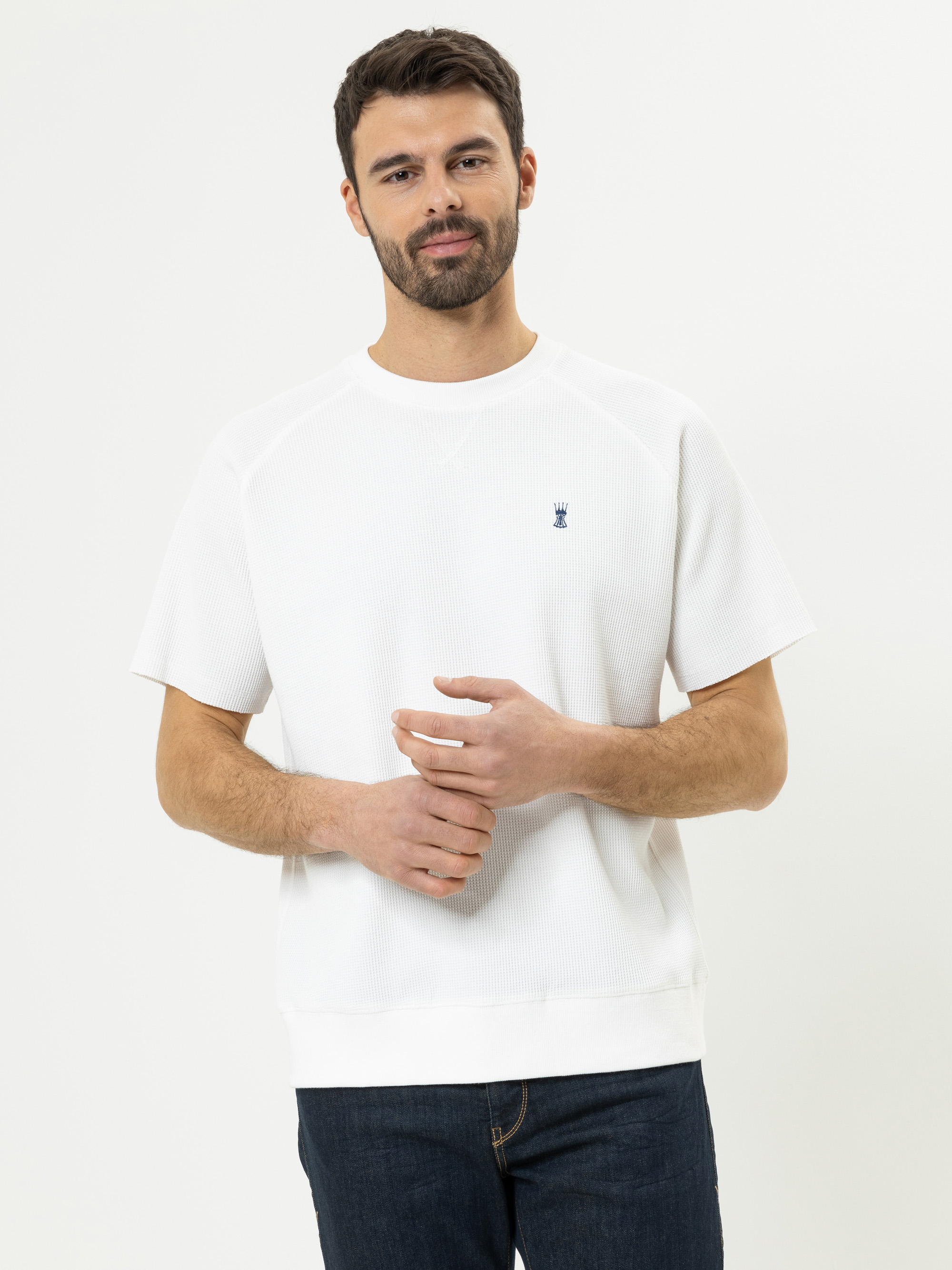 Ein Mann mit kurzen dunklen Haaren und einem Bart trägt ein weißes kurzärmeliges Hemd mit einem kleinen blauen Logo auf der Brust und dunkle Jeans und steht vor einem schlichten weißen Hintergrund.