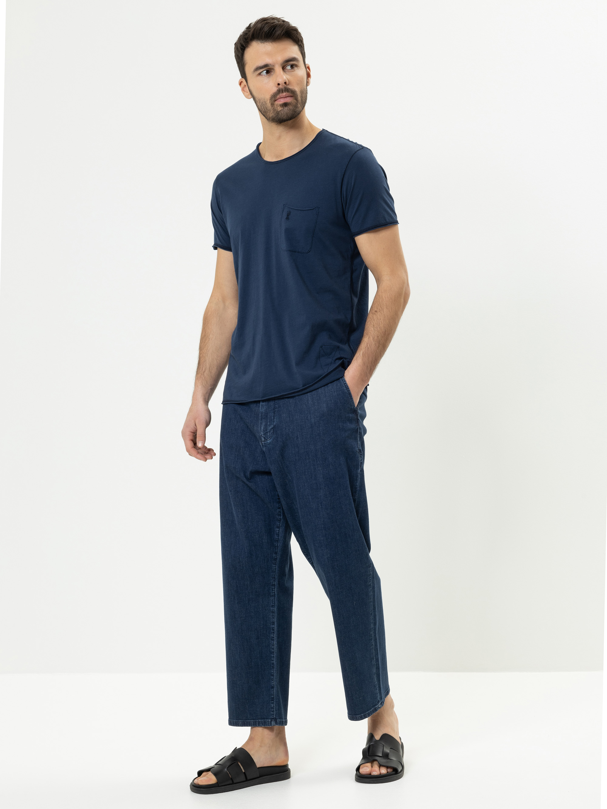 Ein Mann steht vor einem schlichten weißen Hintergrund. Er trägt ein marineblaues T-Shirt mit einer Brusttasche, eine locker sitzende Bluejeans und schwarze Sandalen. Er hat kurzes dunkles Haar und einen Bart und blickt leicht nach links.