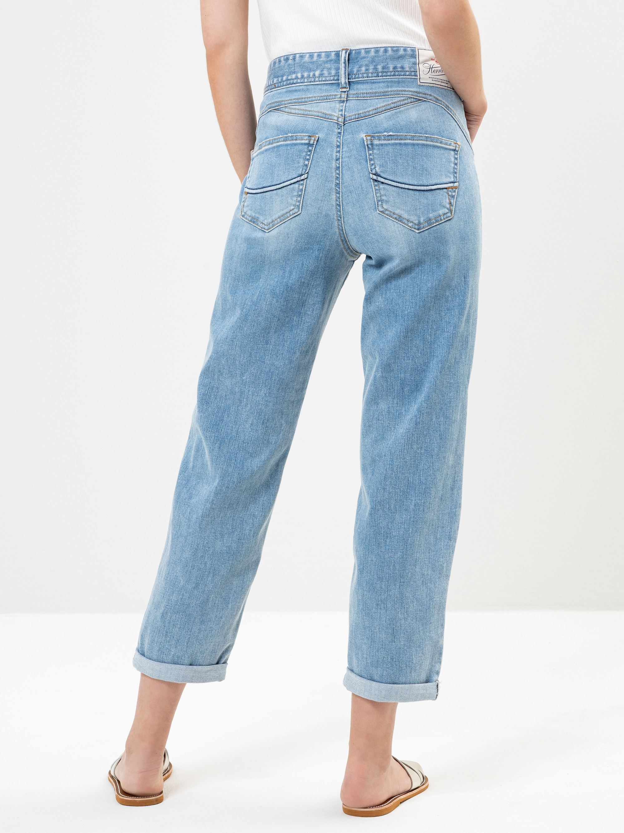 Eine Person steht mit dem Rücken zur Kamera und trägt eine hellblaue Jeans mit hoher Taille und geradem Bein sowie Sandalen mit offenen Zehen. Die Jeans hat zwei Gesäßtaschen und aufgerollte Manschetten. Der Hintergrund ist einfarbig weiß.
