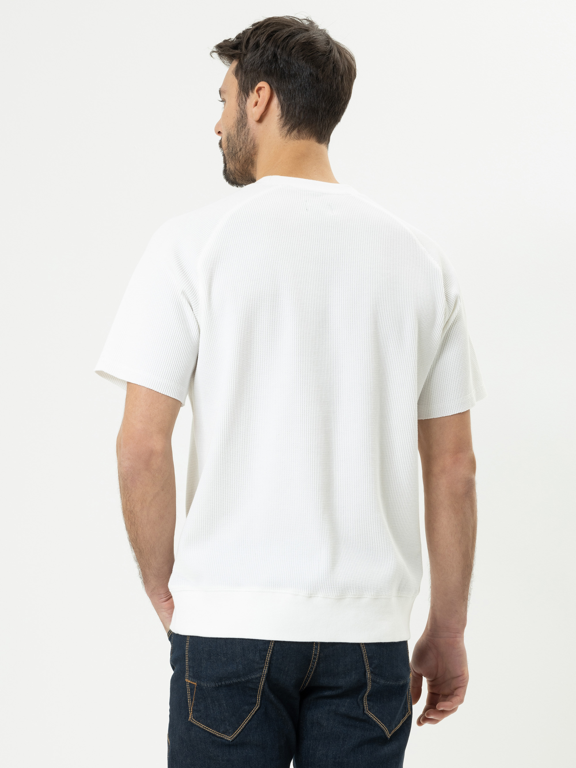 Ein Mann mit kurzen braunen Haaren steht mit dem Rücken zur Kamera, trägt ein weißes kurzärmeliges Hemd und dunkle Jeans vor einem schlichten weißen Hintergrund.