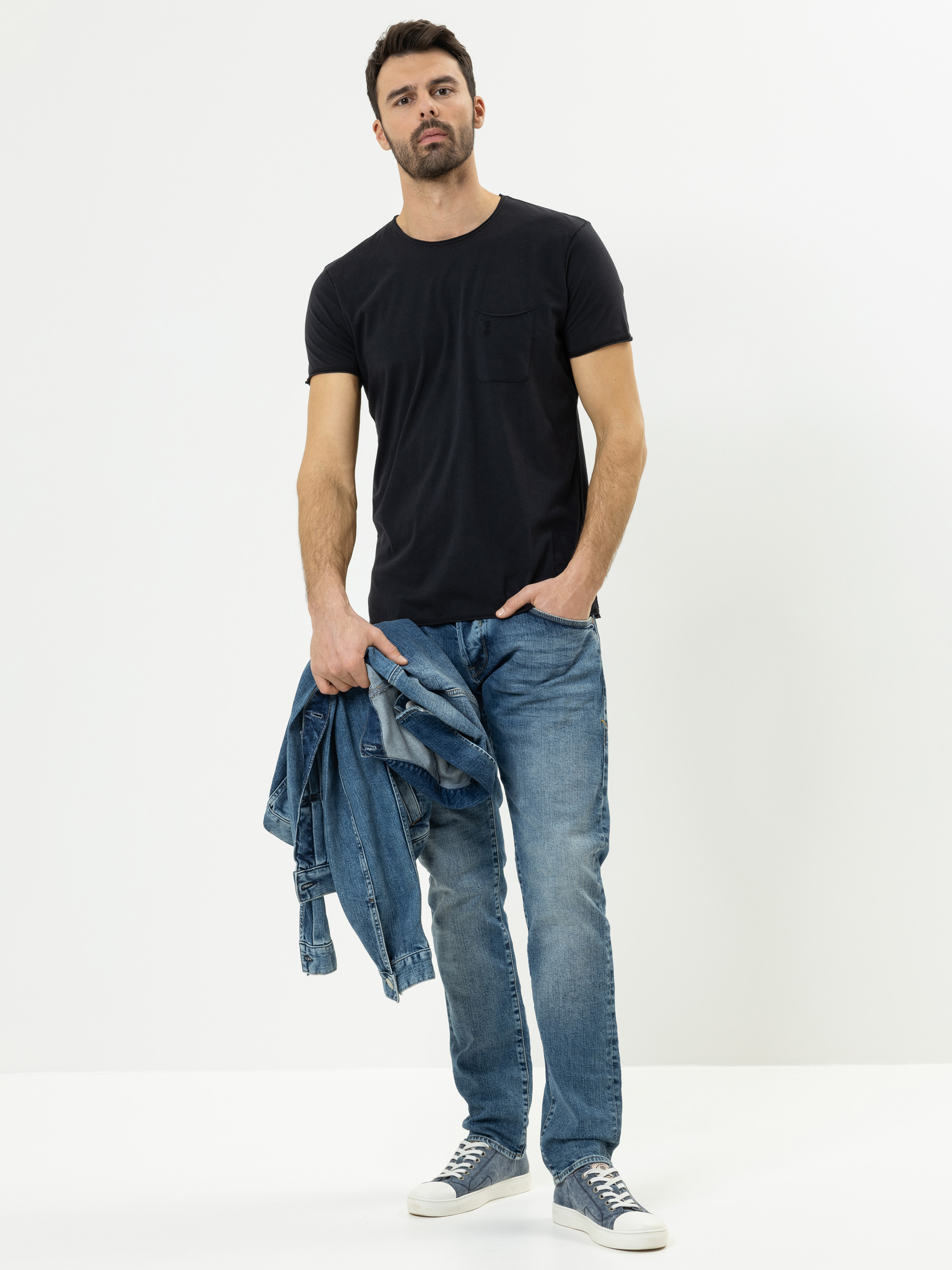 Ein Mann mit kurzen dunklen Haaren und einem Bart trägt ein schwarzes T-Shirt, blaue Jeans und graue Turnschuhe. Er hält eine blaue Jeansjacke in seiner linken Hand und steht vor einem schlichten weißen Hintergrund.