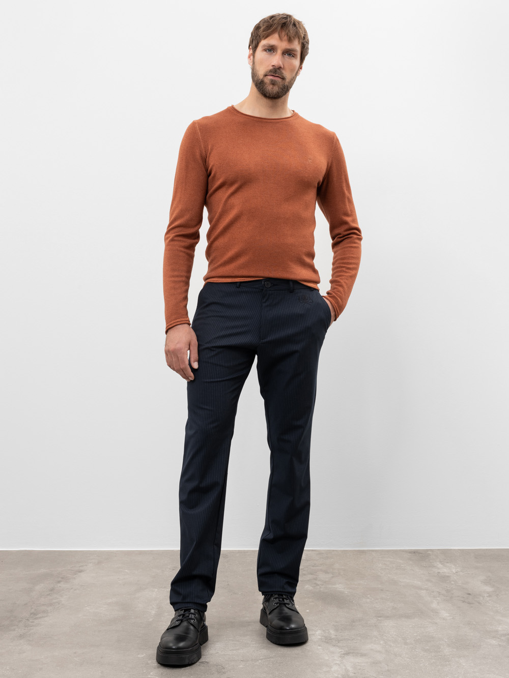 Vor einem schlichten weißen Hintergrund steht ein Mann mit kurzen Haaren und Bart, der ein rostfarbenes langärmeliges Hemd, eine dunkelblaue Hose und schwarze Schuhe trägt und eine Hand in der Tasche hat.