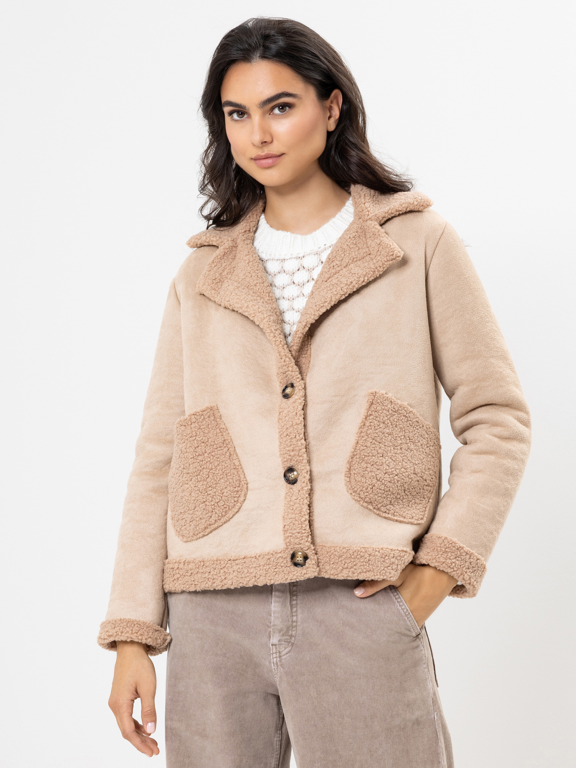 Eine Frau, die eine beigefarbene Sherpa-Jacke mit großen Vordertaschen über einem weißen Strickoberteil und einer taupefarbenen Hose trägt, steht vor einem schlichten weißen Hintergrund.