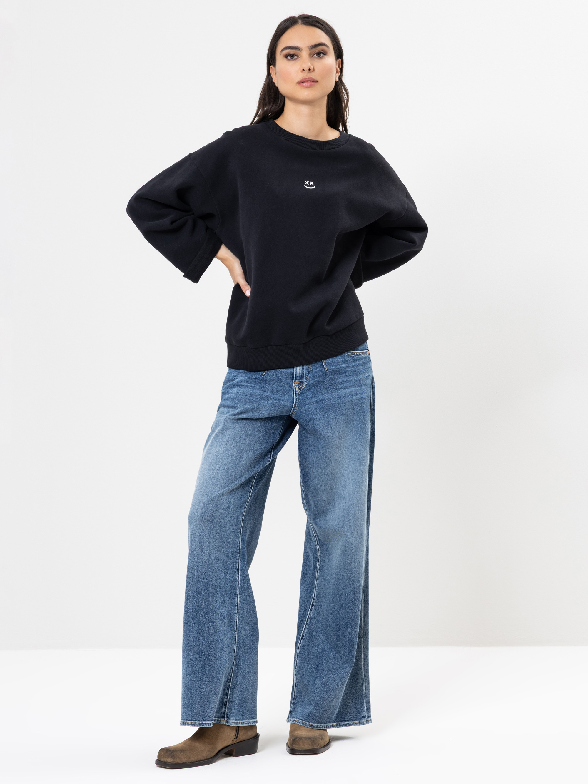 Eine Frau steht vor einem schlichten weißen Hintergrund. Sie trägt ein lockeres schwarzes Sweatshirt, weit geschnittene Bluejeans und braune Schuhe. Ihre Hände ruhen auf den Hüften, und sie hat einen neutralen Gesichtsausdruck und lange dunkle Haare.