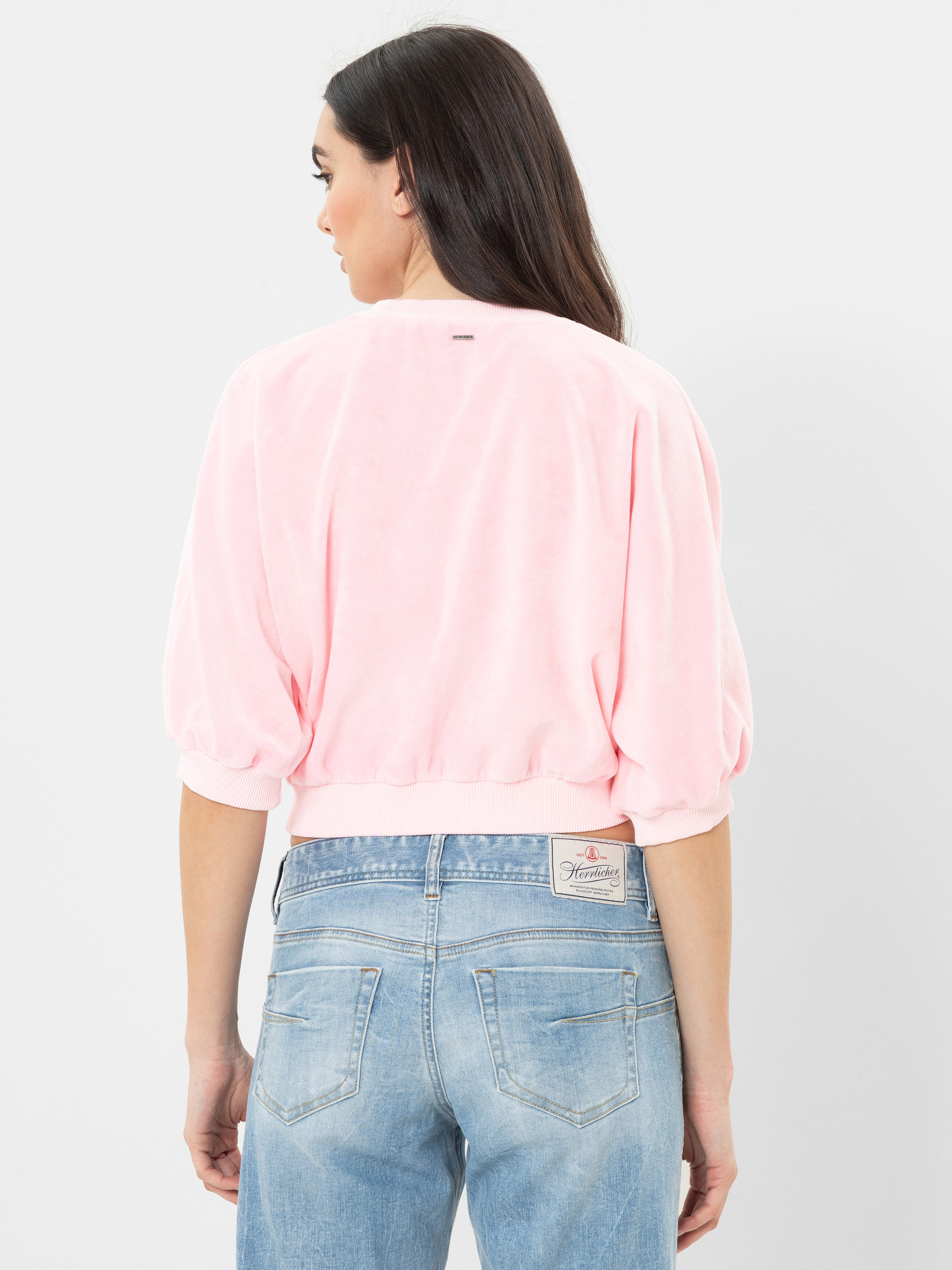 Frau mit langen dunklen Haaren, bekleidet mit einem hellrosa, locker sitzenden Oberteil mit ellbogenlangen Ärmeln und blauen Jeans mit hoher Taille, von hinten gesehen, vor einem schlichten weißen Hintergrund.