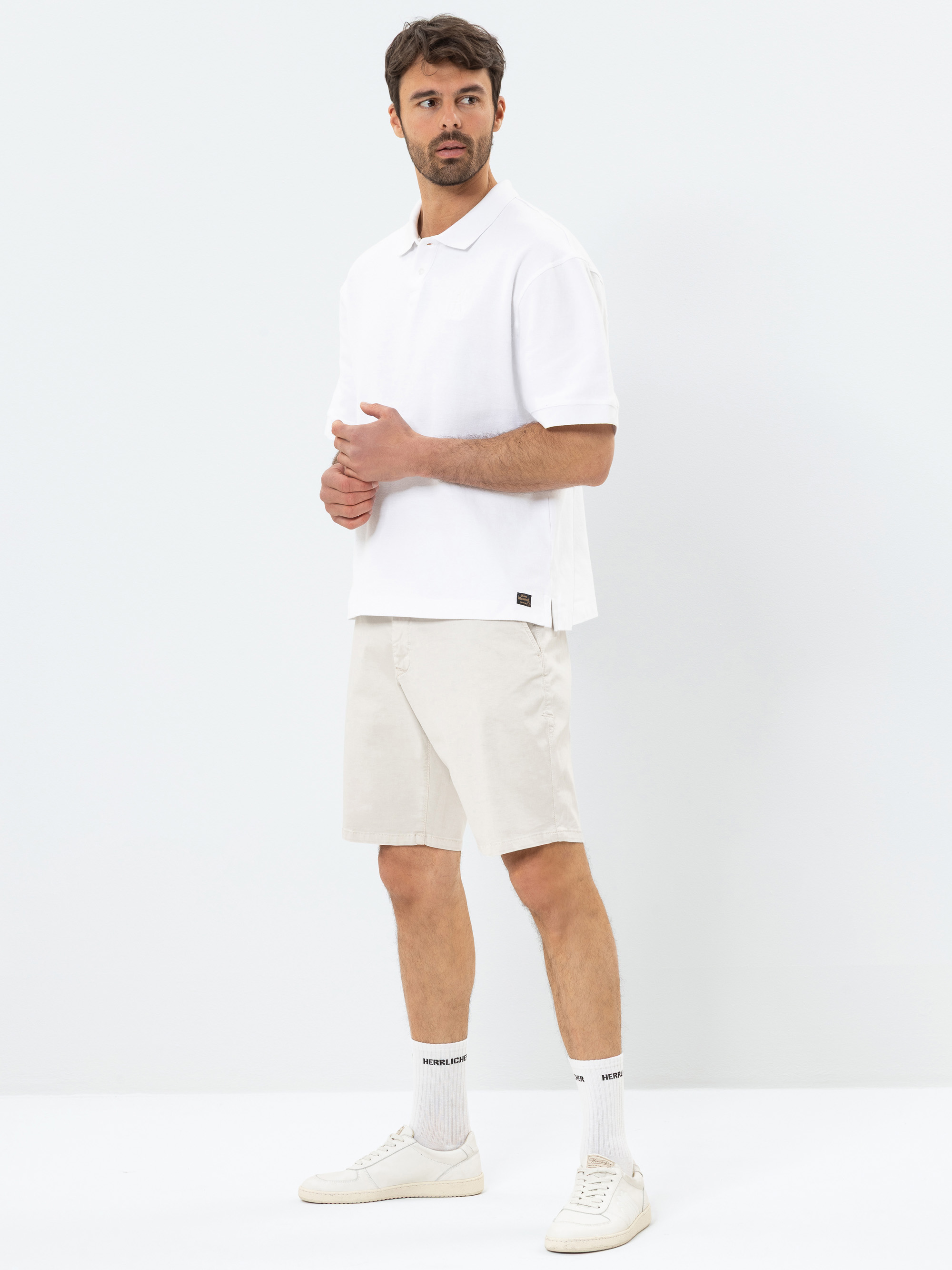 Ein Mann steht vor einem schlichten weißen Hintergrund und trägt ein weißes Polohemd, hellbeige Shorts, weiße Socken mit schwarzem Text und weiße Turnschuhe. Er blickt leicht zur Seite und stützt eine Hand auf seinen Oberkörper.