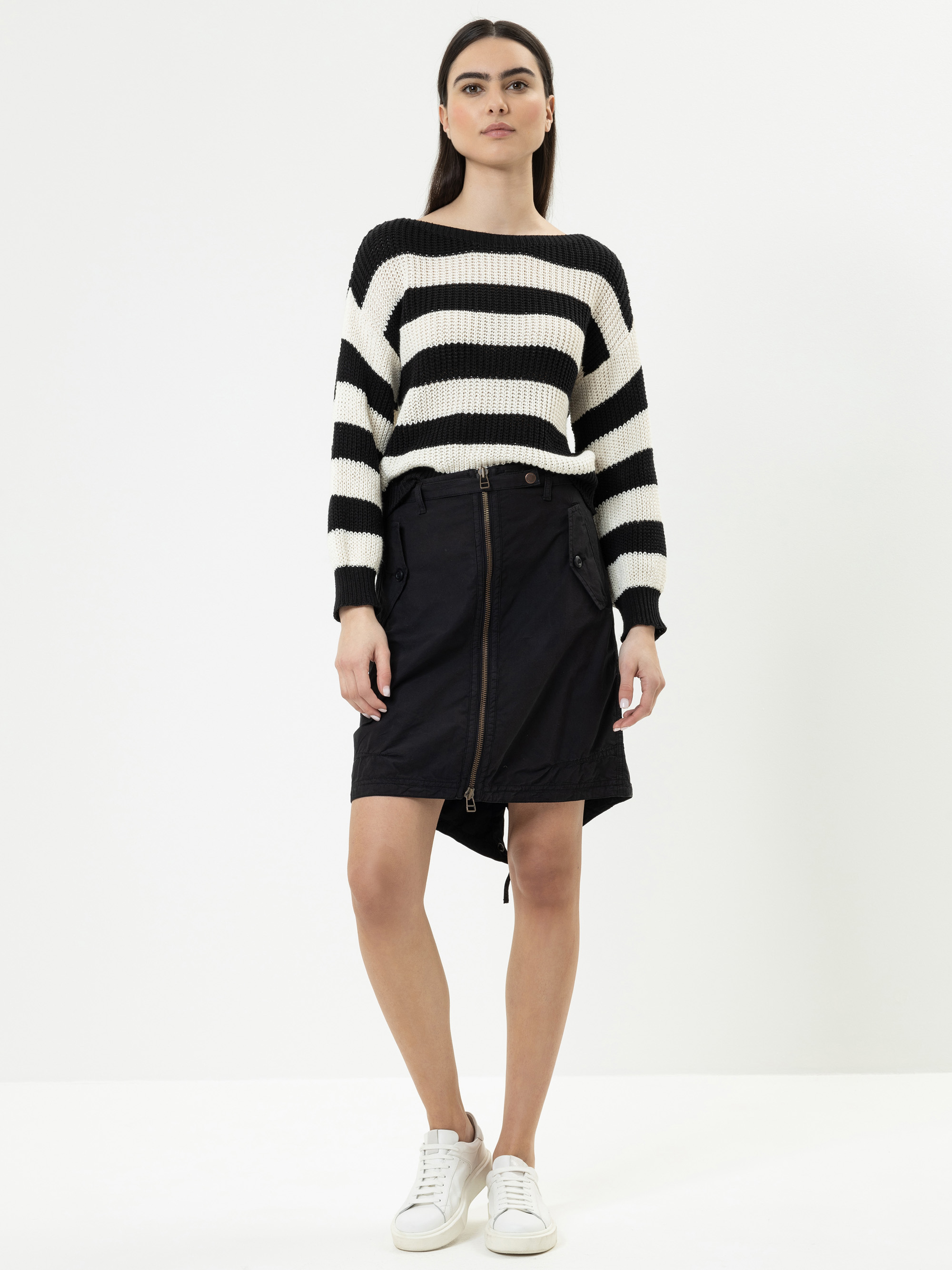 Eine Frau steht vor einem weißen Hintergrund und trägt einen schwarz-weiß gestreiften Pullover, einen schwarzen Rock mit Reißverschluss und weiße Turnschuhe. Sie hat glattes dunkles Haar und einen neutralen Gesichtsausdruck.