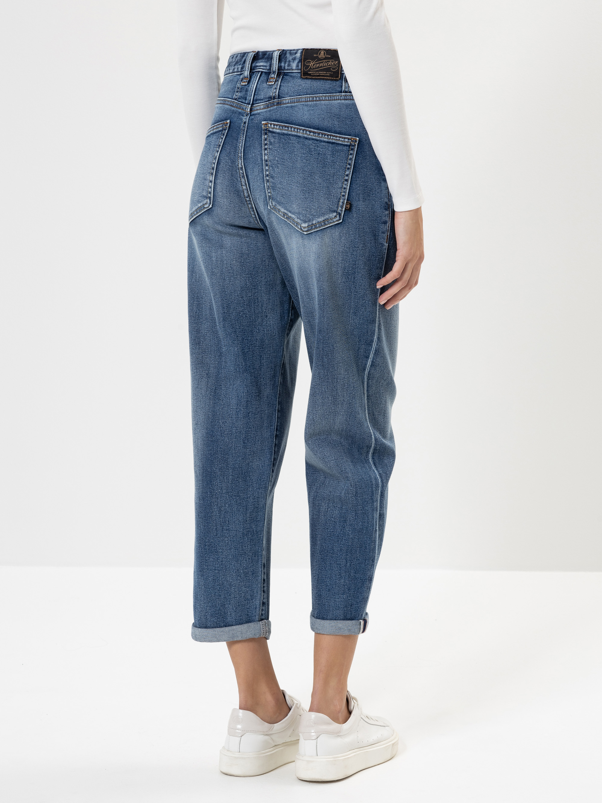 Eine Person steht mit dem Rücken zur Kamera, trägt eine blaue Jeans mit Bündchen, ein weißes langärmeliges Oberteil und weiße Turnschuhe vor einem schlichten weißen Hintergrund.