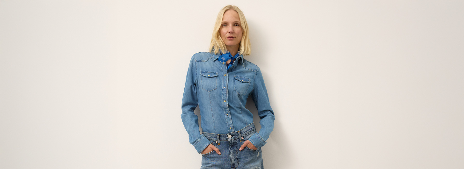 Eine Frau mit blondem Haar steht vor einer schlichten weißen Wand. Sie trägt ein Jeanshemd, dazu passende Jeans und einen blau gemusterten Schal um den Hals, die Hände in den Taschen.