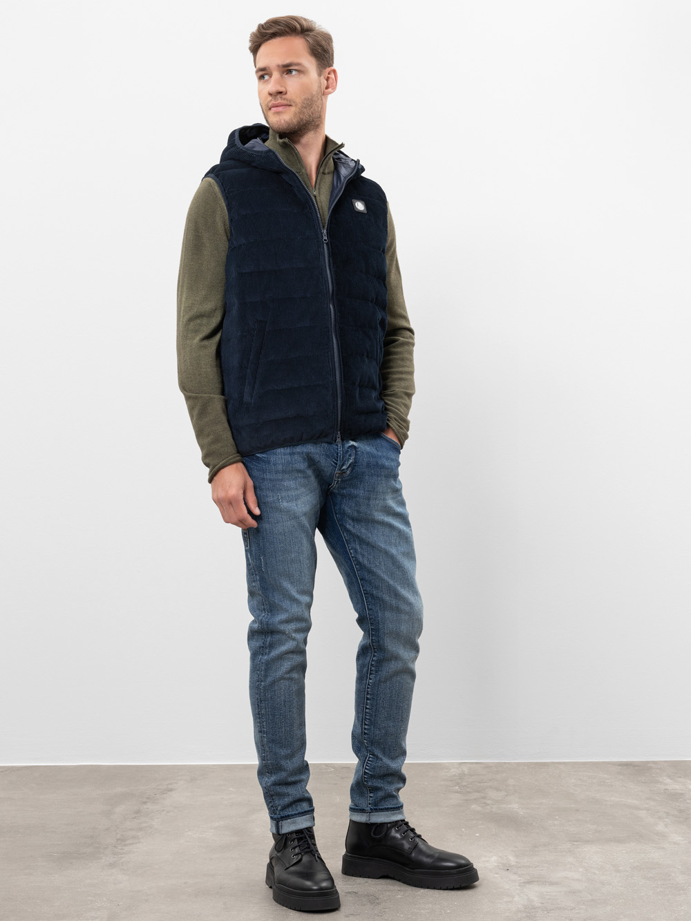 Ein Mann steht in einem Haus und trägt eine dunkelblaue Kapuzenweste über einem olivgrünen Pullover mit Reißverschluss, blaue Jeans und schwarze Stiefel vor einer schlichten weißen Wand und einem grauen Boden.