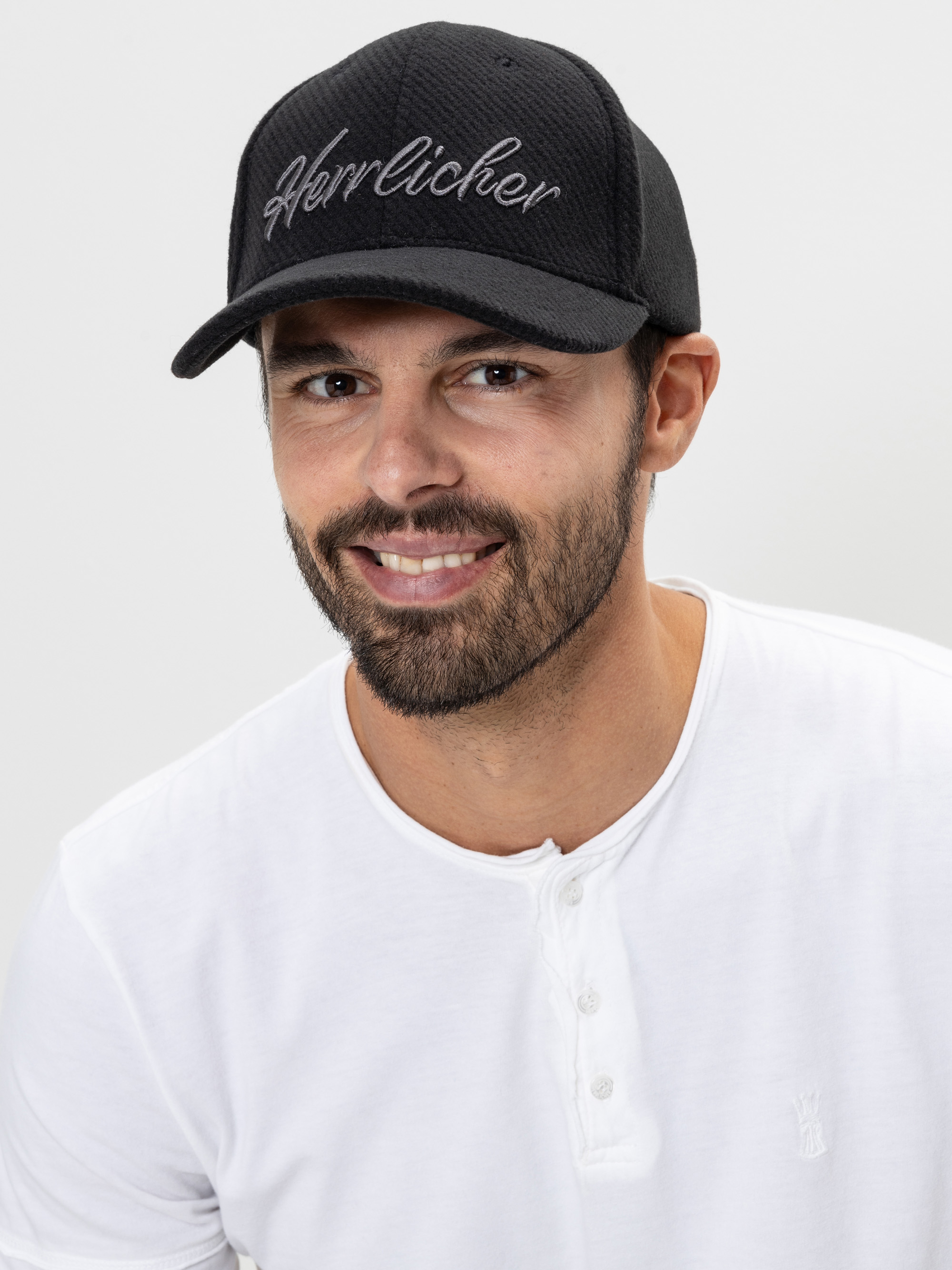 Ein Mann mit kurzem Bart und Schnurrbart, der eine schwarze Mütze mit dem aufgestickten Wort Herrlicher und ein weißes Hemd trägt, lächelt in die Kamera vor einem schlichten hellen Hintergrund.