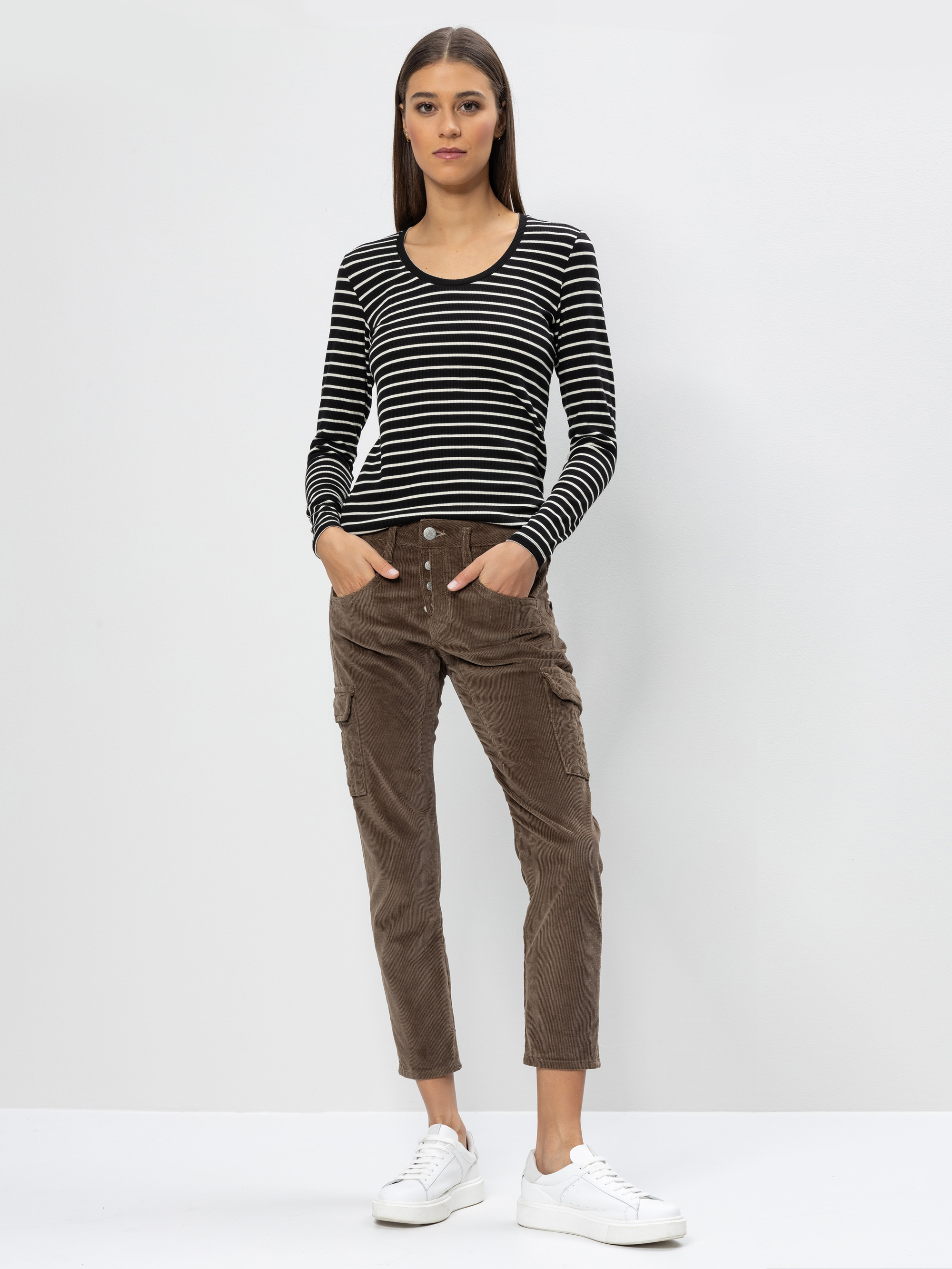 Eine Frau steht vor einem schlichten, hellen Hintergrund und trägt ein schwarz-weiß gestreiftes, langärmeliges Oberteil, eine braune Cordhose mit Seitentaschen und weiße Turnschuhe. Ihre Hände stecken in den Taschen, und sie hat glattes braunes Haar.