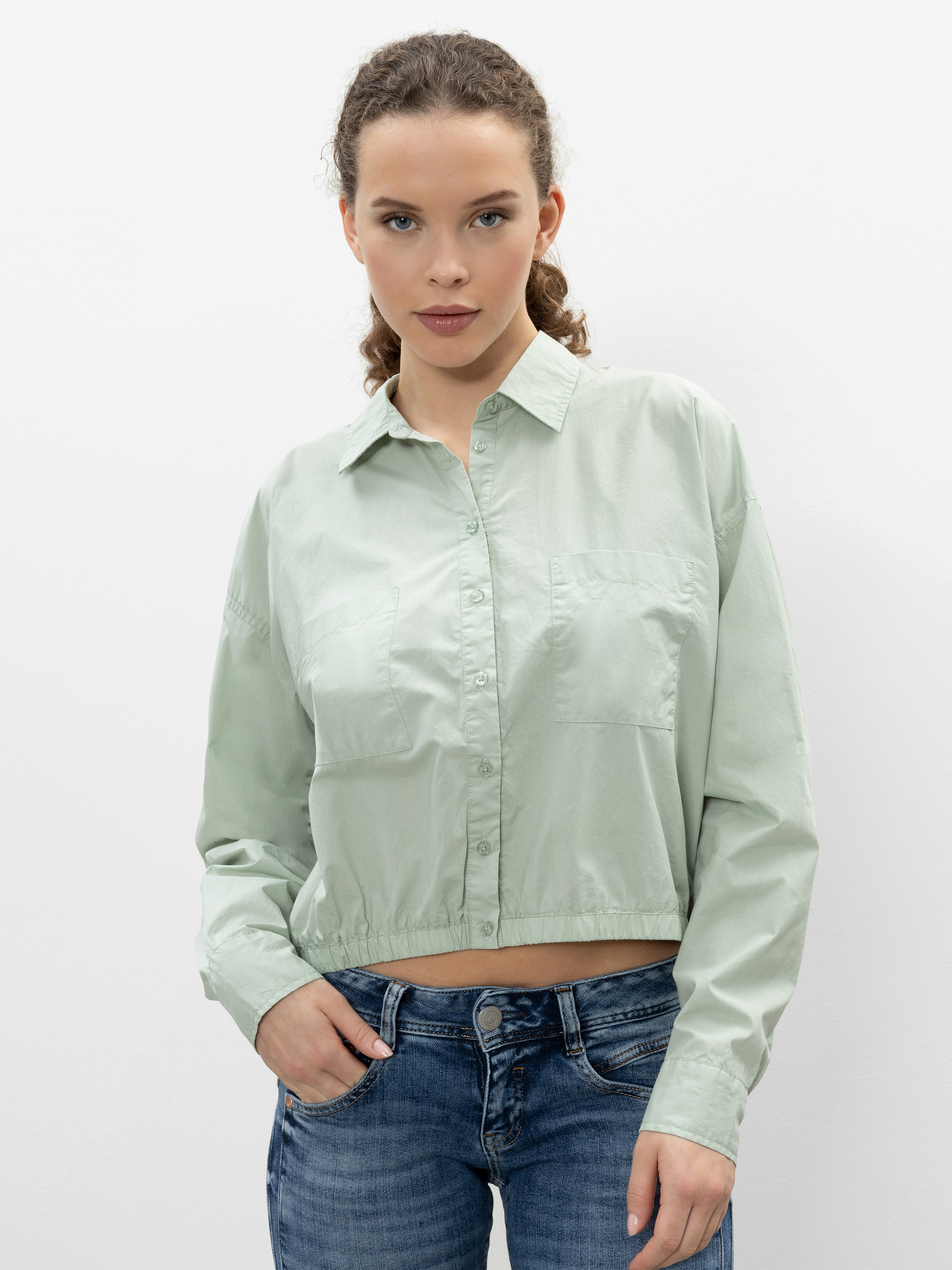 Eine Frau mit lockigem Haar, die ein hellgrünes, langärmeliges Button-up-Shirt und blaue Jeans trägt, steht vor einem schlichten weißen Hintergrund und blickt selbstbewusst in die Kamera.