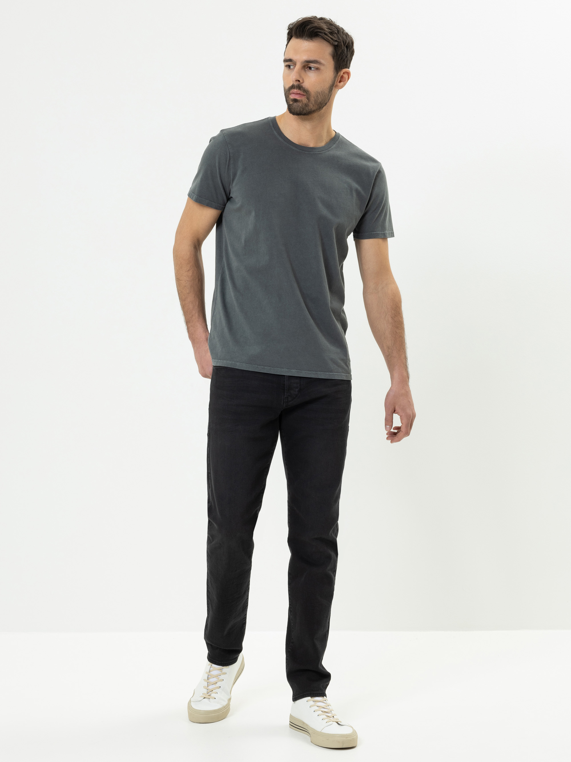 Ein Mann mit kurzen dunklen Haaren, Bart und Schnurrbart trägt ein schlichtes graues T-Shirt, schwarze Jeans und weiße Turnschuhe und steht mit entspannter Körperhaltung vor einem schlichten weißen Hintergrund.