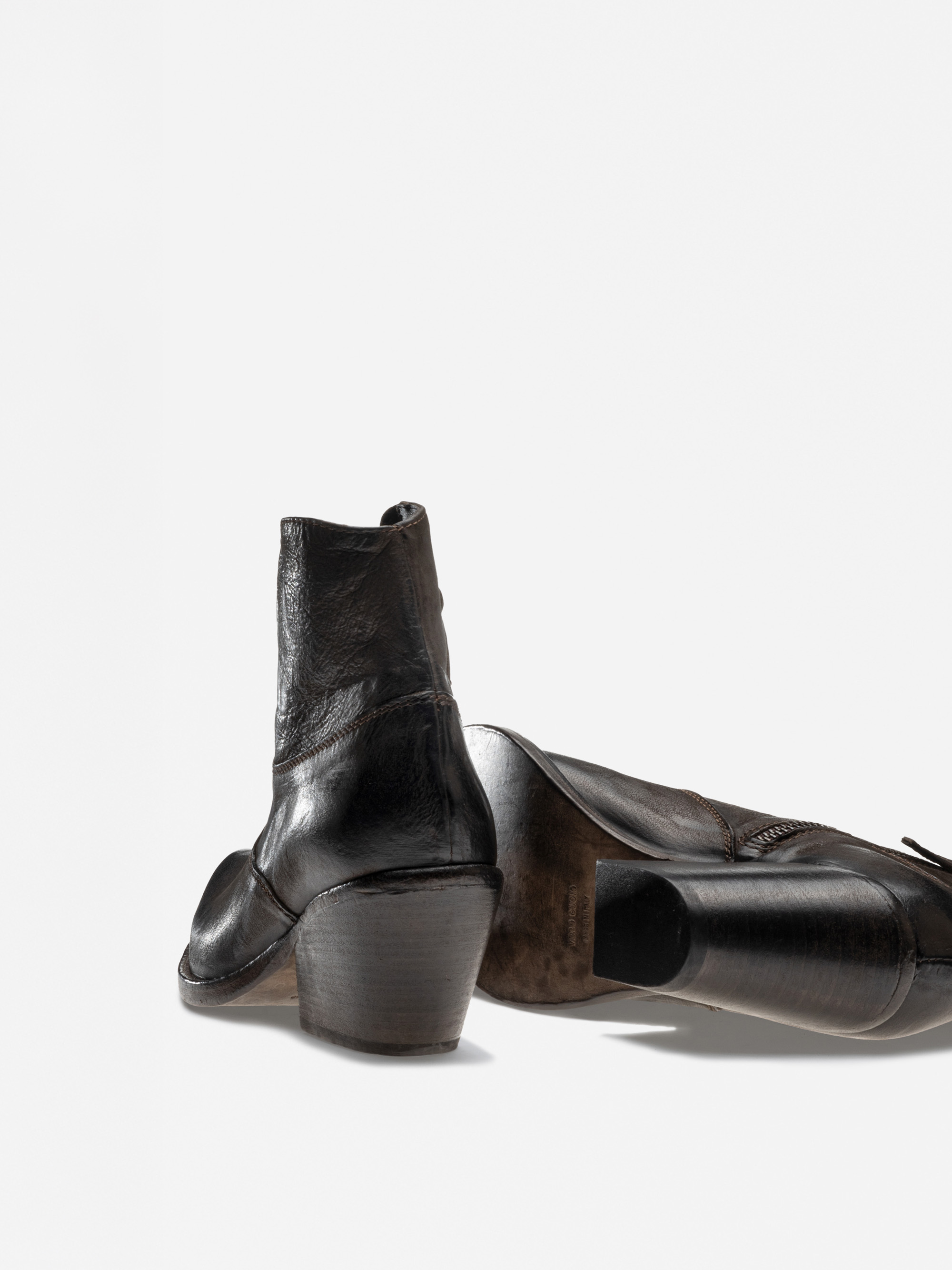 Ein Paar schwarze Lederstiefel mit klobigen Holzabsätzen. Ein Stiefel steht aufrecht, während der andere auf der Seite liegt und die abgenutzte Sohle zeigt. Der Hintergrund ist schlicht und weiß.