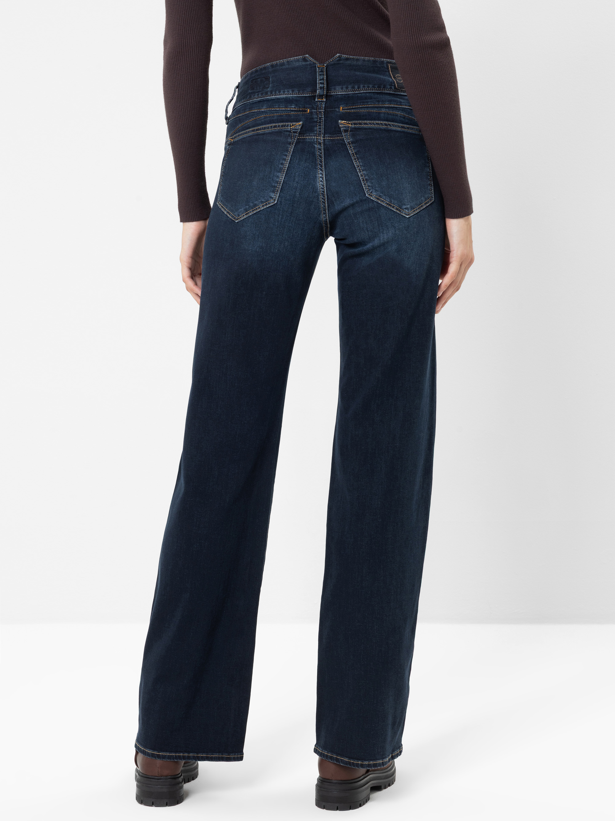 Eine Person, die eine dunkelblaue Jeans mit hoher Taille und weitem Bein trägt, von hinten gesehen. Die Jeans hat zwei Gesäßtaschen und gelbe Nähte. Die Person trägt außerdem ein langärmeliges, dunkles Oberteil und schwarze Schuhe.