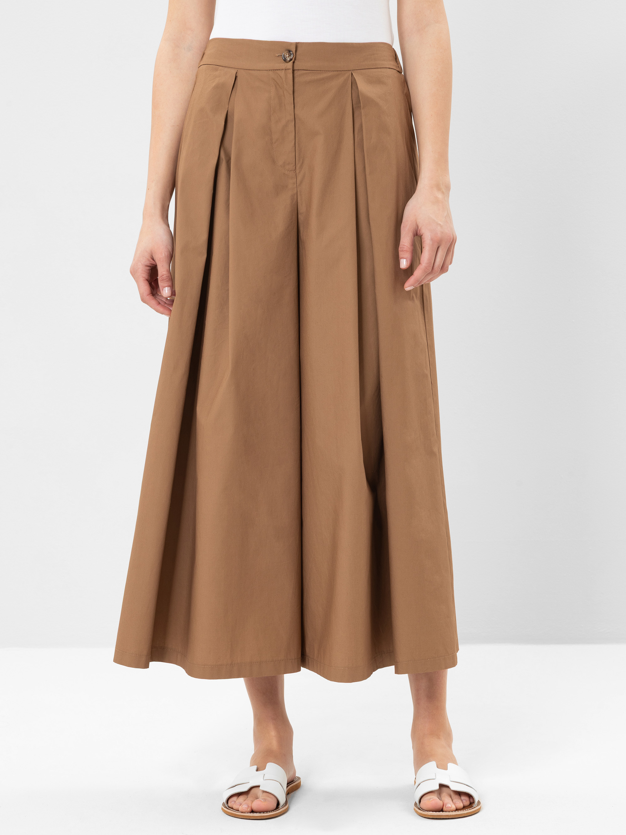 Eine Person, die ein weißes ärmelloses Oberteil, eine hellbraune Culottes mit weitem Bein und weiße Sandalen trägt, steht vor einem schlichten hellen Hintergrund.
