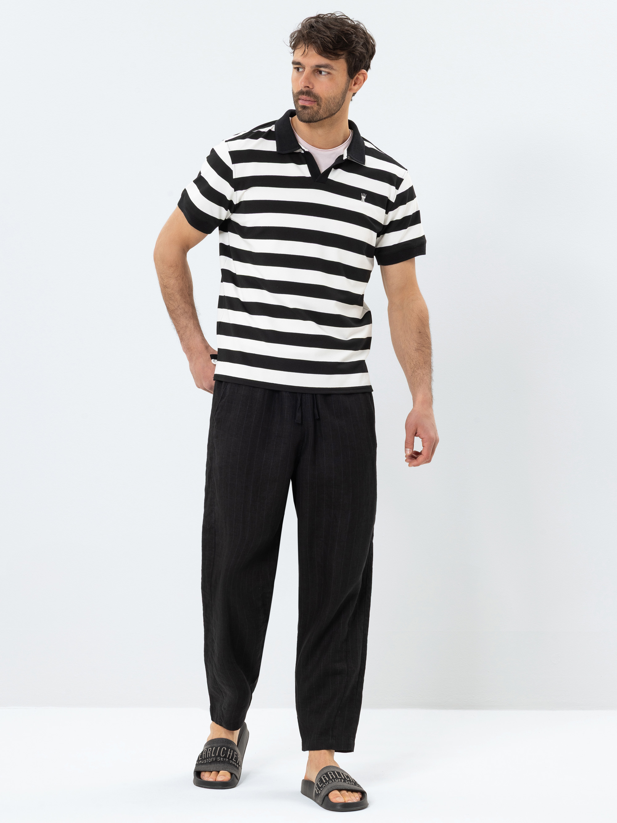 Ein Mann steht vor einem schlichten Hintergrund und trägt ein schwarz-weiß gestreiftes Poloshirt, eine weite schwarze Hose und graue Slide-Sandalen. Er hat kurzes, gewelltes Haar und einen Bart, und seine rechte Hand steckt in der Tasche.
