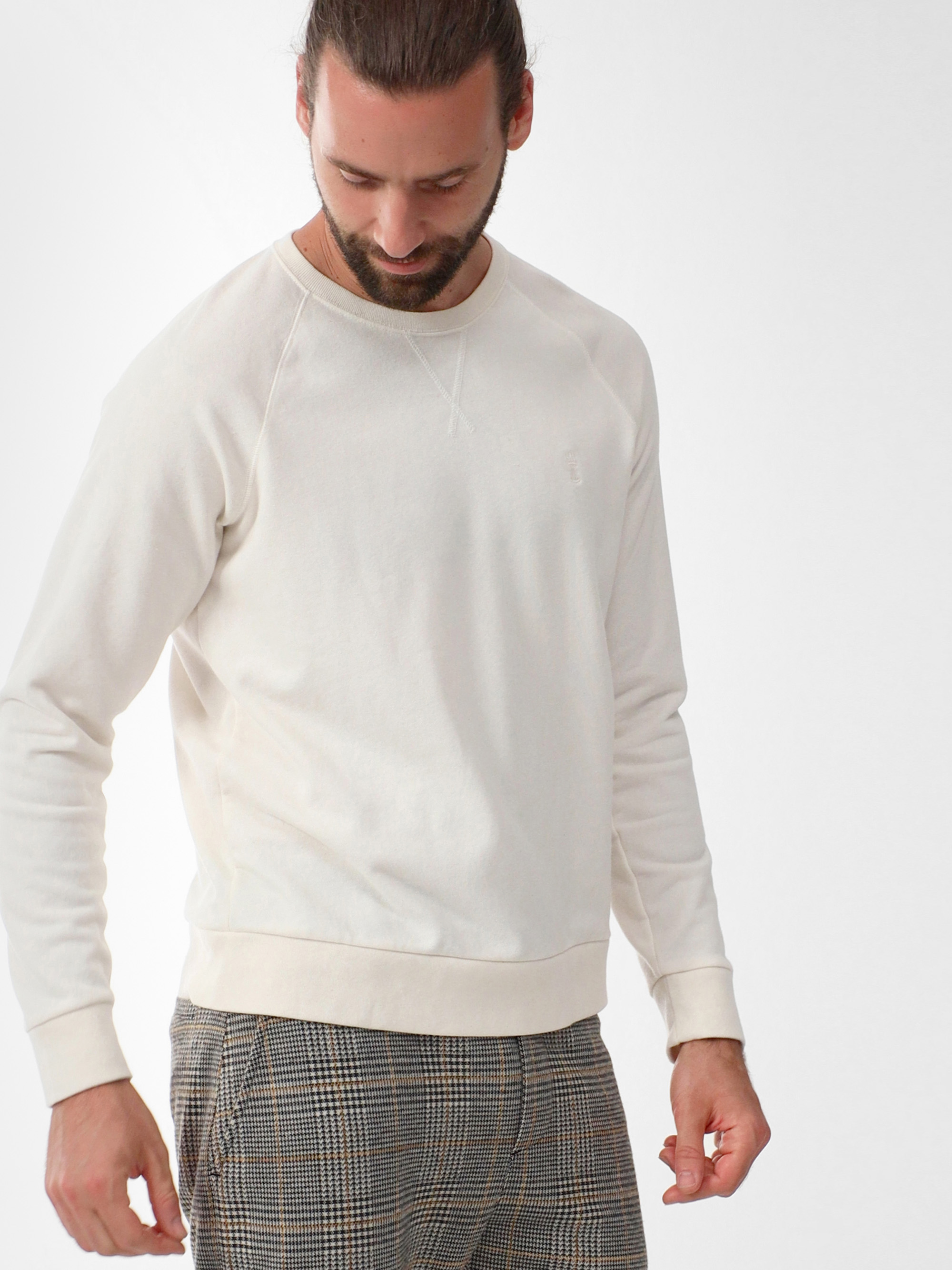 Ein Mann mit langen, nach hinten gebundenen Haaren trägt ein cremefarbenes Sweatshirt und eine karierte Hose und blickt mit einem leichten Lächeln auf einen schlichten weißen Hintergrund.