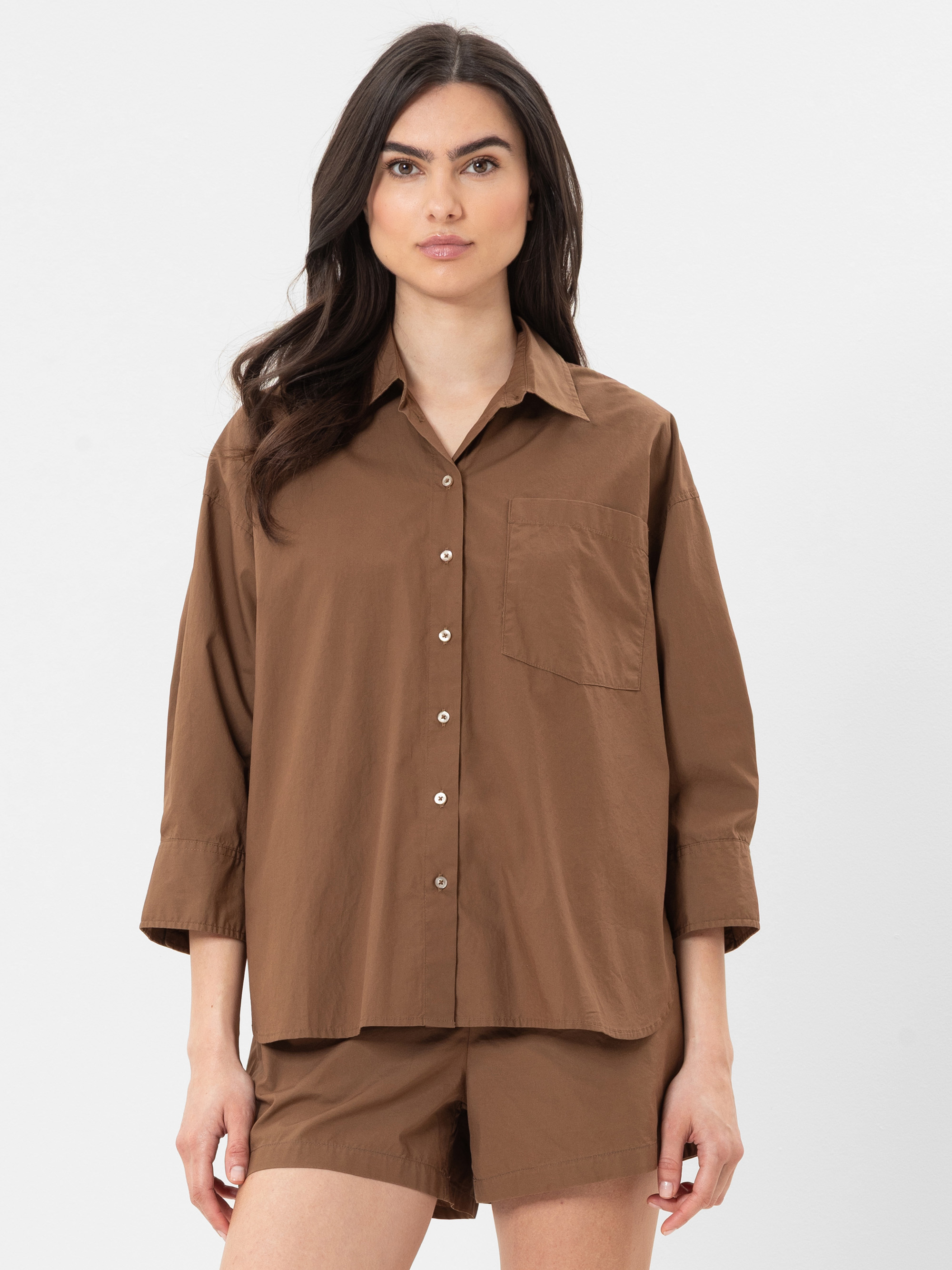 Eine Frau, die ein locker sitzendes braunes Button-up-Hemd mit Dreiviertelärmeln und passende braune Shorts trägt, steht vor einem schlichten weißen Hintergrund.
