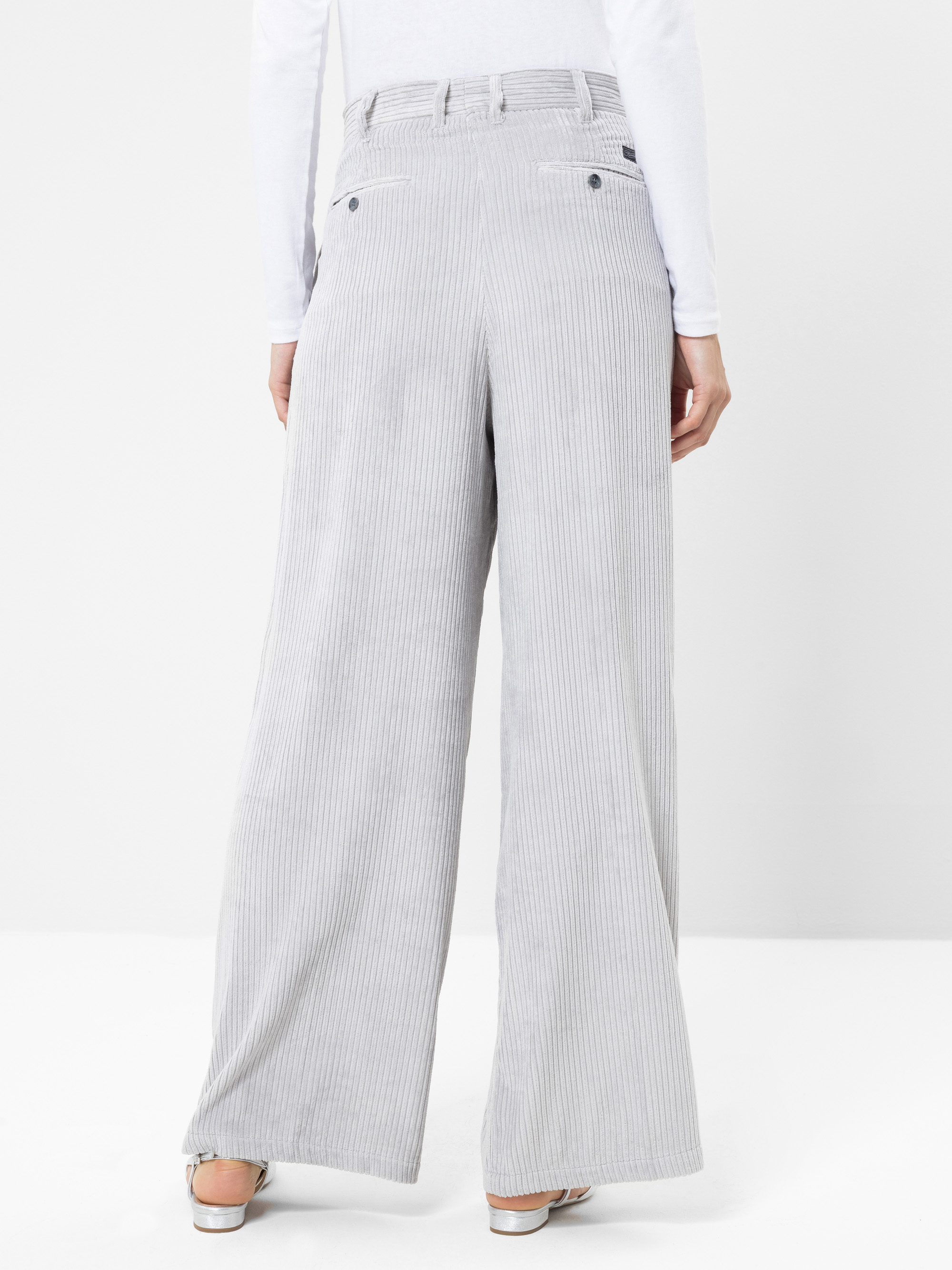 Eine Person, die eine hellgraue Cordhose mit hoher Taille und weitem Bein sowie ein weißes Langarm-Oberteil trägt, von hinten vor einem schlichten weißen Hintergrund gesehen.