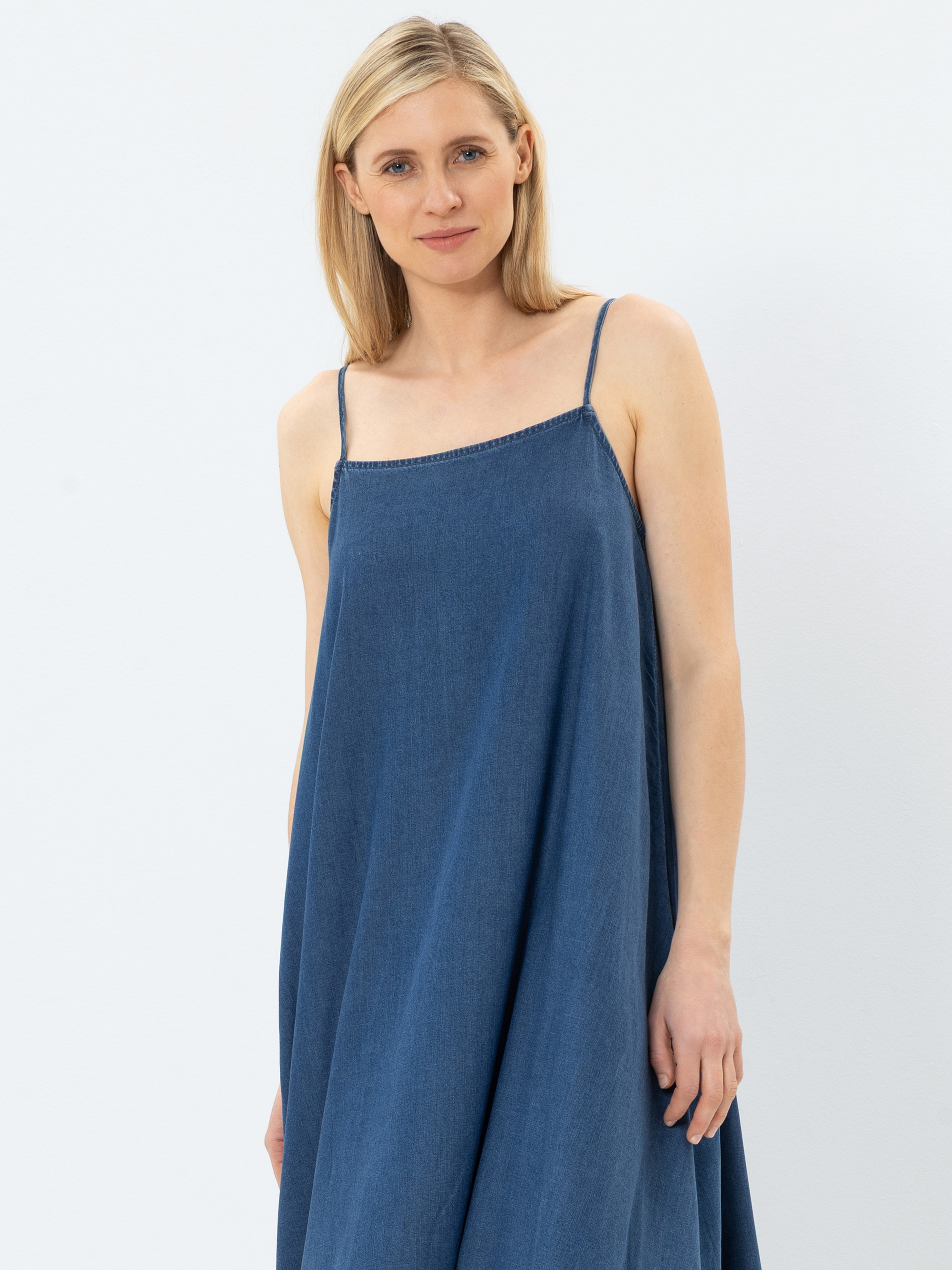 Eine Frau mit glattem, schulterlangem blondem Haar trägt ein locker sitzendes, ärmelloses blaues Kleid mit Spaghetti-Trägern, steht vor einem schlichten weißen Hintergrund und lächelt sanft.