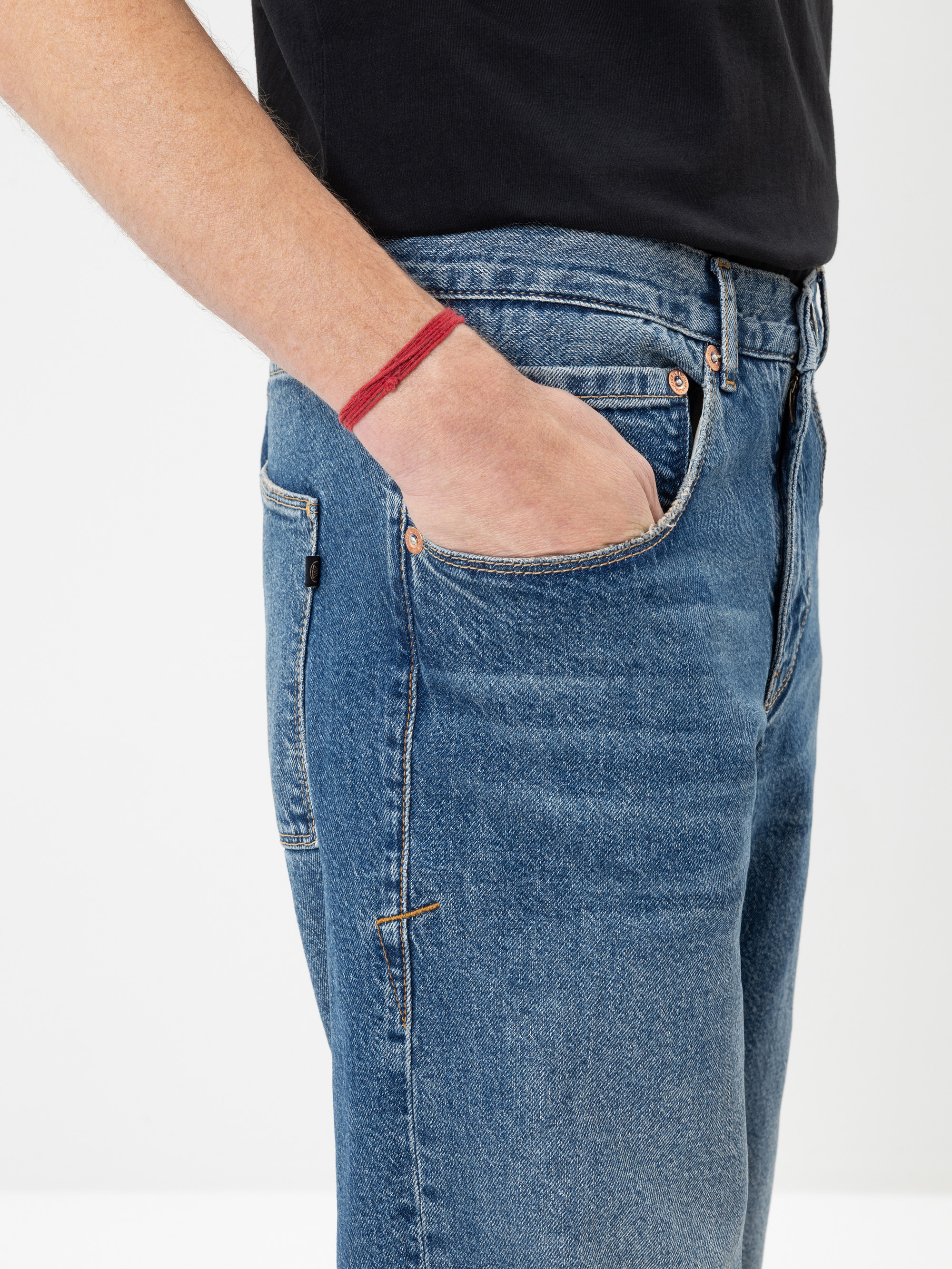 Eine Person, die blaue Jeans und ein schwarzes Hemd trägt, steht da und hat die linke Hand in der Tasche. Am Handgelenk ist ein rotes Armband oder ein rotes Zeichen zu sehen. Der Hintergrund ist einfarbig und weiß.