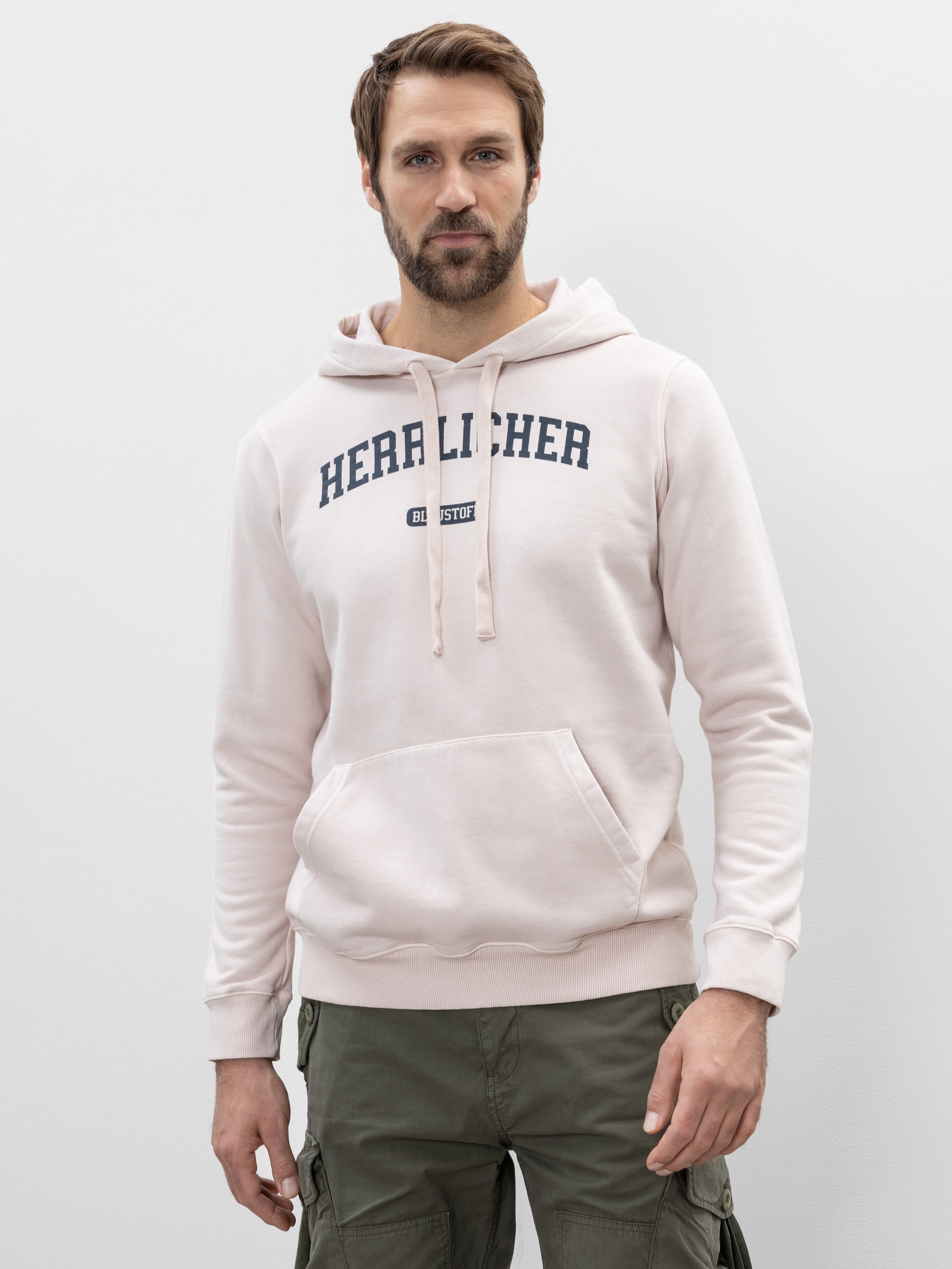 Ein Mann mit kurzen braunen Haaren und einem Bart trägt ein hellrosa Kapuzensweatshirt mit dem Aufdruck HERAL LICHER in dunklen Buchstaben und eine olivgrüne Cargohose vor einem schlichten hellen Hintergrund.