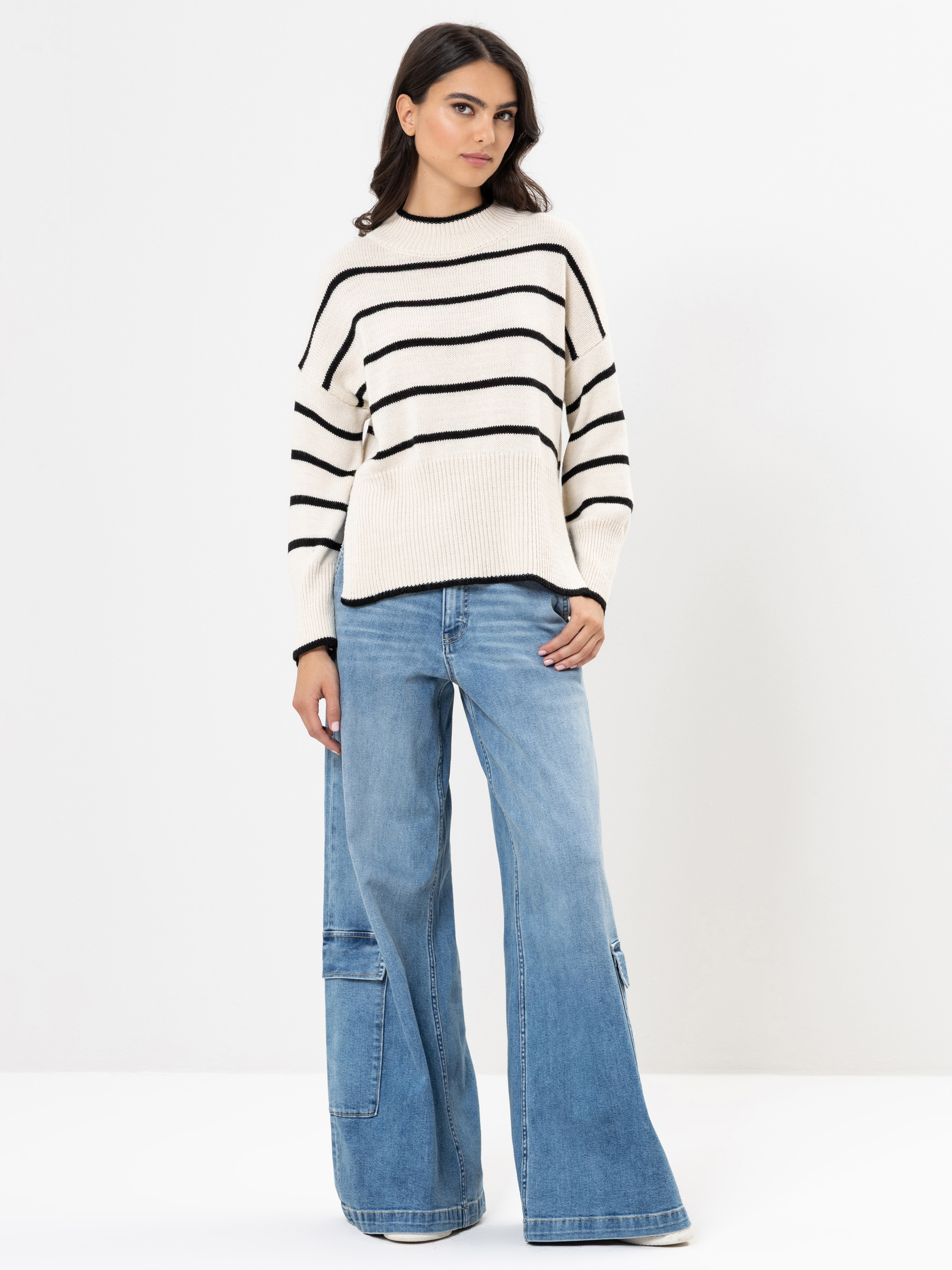 Eine Frau steht vor einem schlichten weißen Hintergrund und trägt einen cremefarbenen Pullover mit schwarzen Querstreifen und eine weit geschnittene blaue Jeans mit aufgesetzten Taschen. Sie hat dunkles Haar und einen entspannten, selbstbewussten Gesich