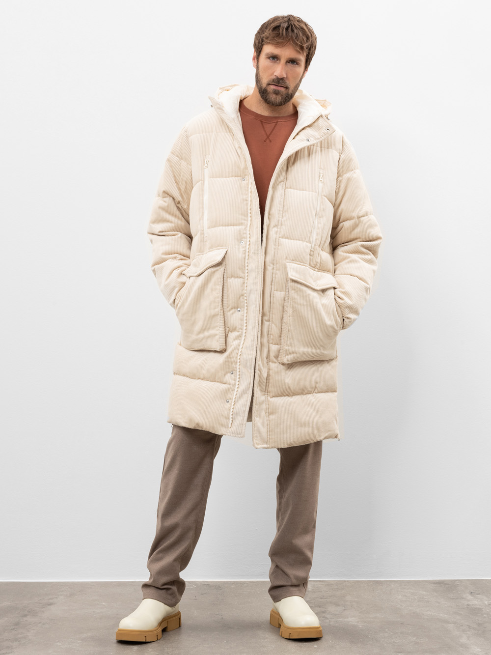 Ein Mann steht vor einem schlichten weißen Hintergrund und trägt einen langen, cremefarbenen wattierten Mantel über einem rostfarbenen Sweatshirt, eine taupefarbene Hose und beigefarbene Slipper. Seine Hände stecken in den Manteltaschen.