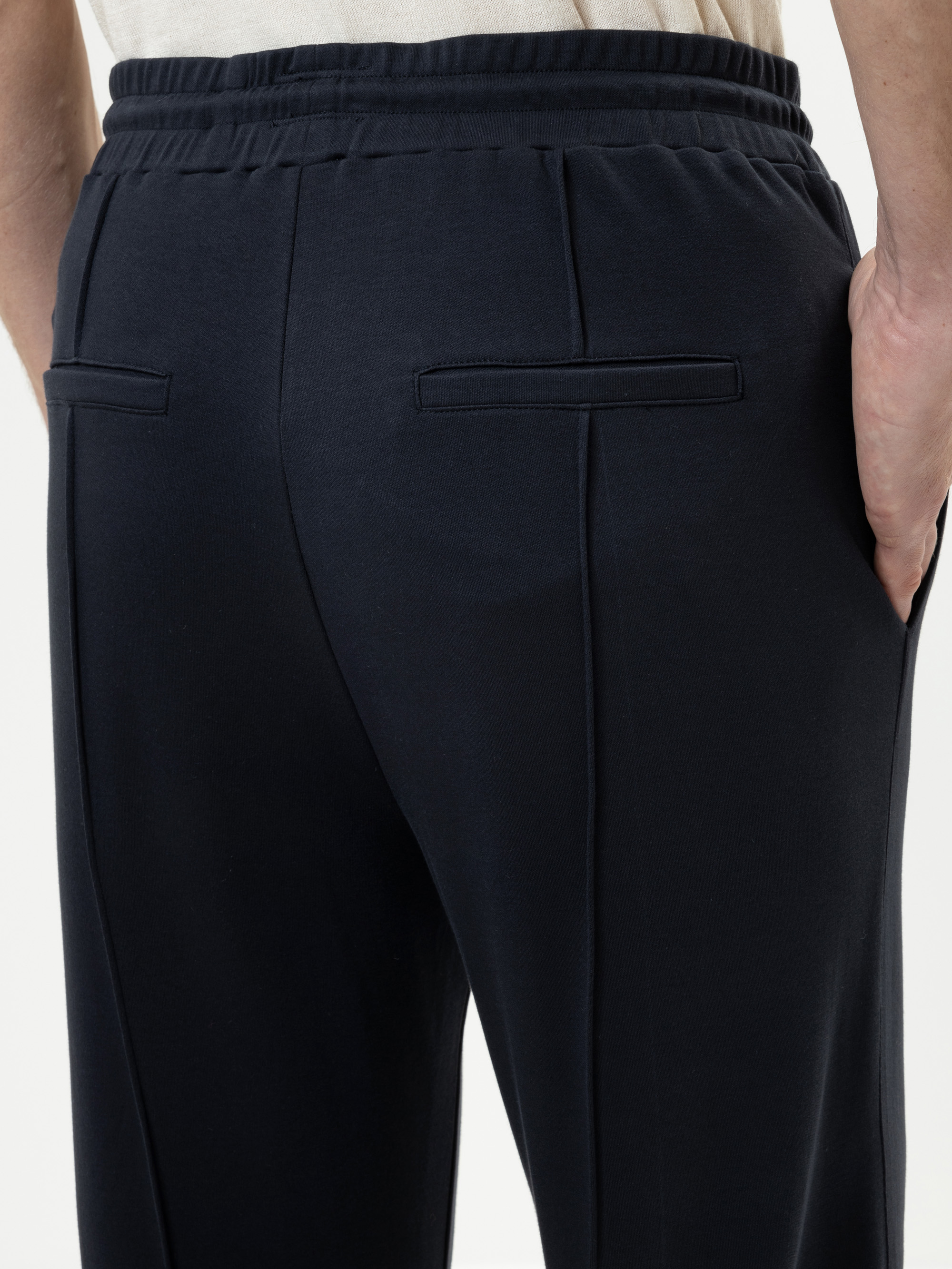 Nahaufnahme einer Person, die eine dunkle marineblaue Hose mit Leistentaschen auf der Rückseite trägt und mit einer Hand in ihrer Tasche steht. Die Hose hat einen elastischen Bund und eine lockere Passform.