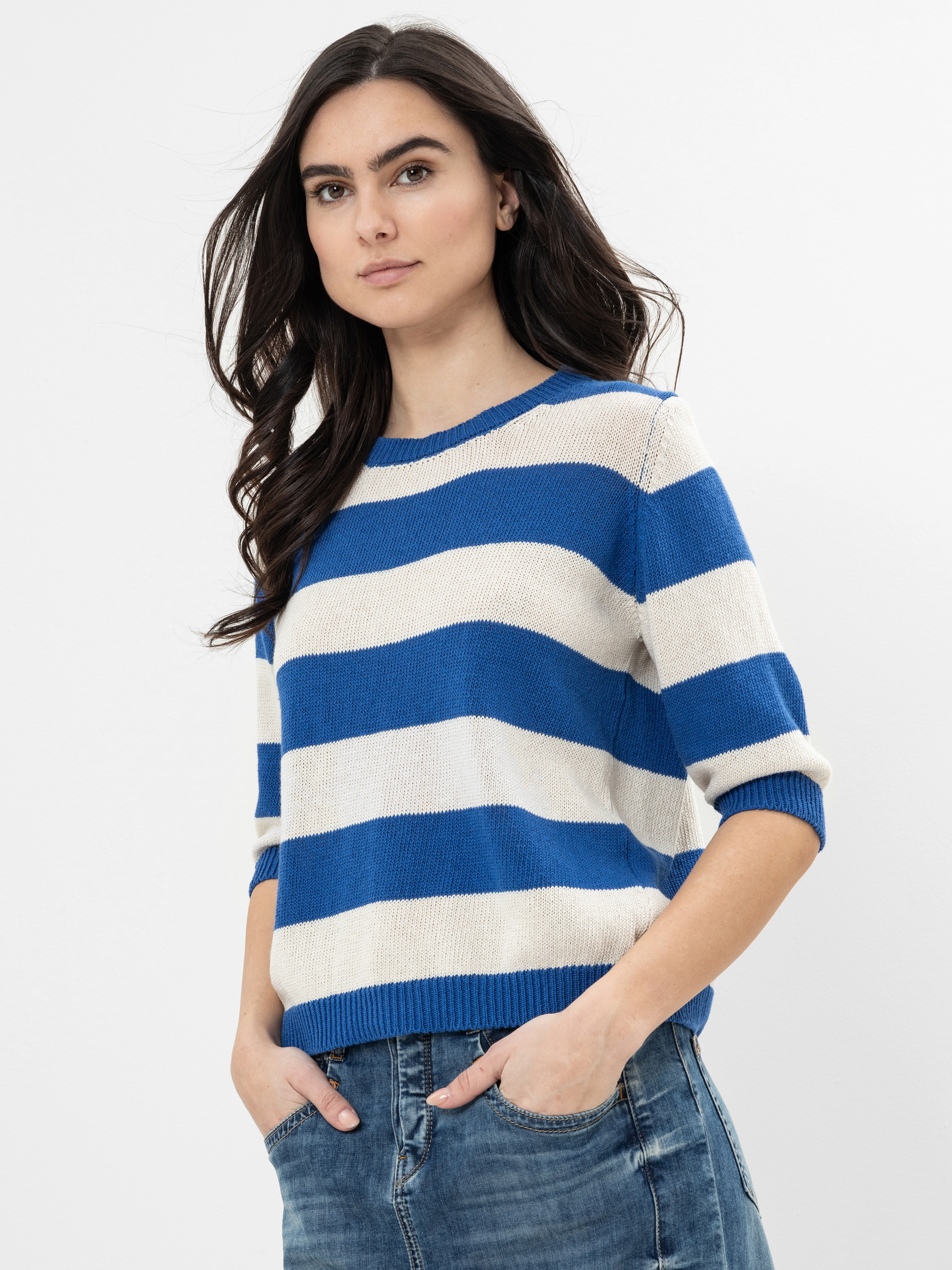 Eine Frau mit langen dunklen Haaren trägt einen blau-weiß gestreiften Pullover mit ellenbogenlangen Ärmeln und blaue Jeans vor einem schlichten weißen Hintergrund.